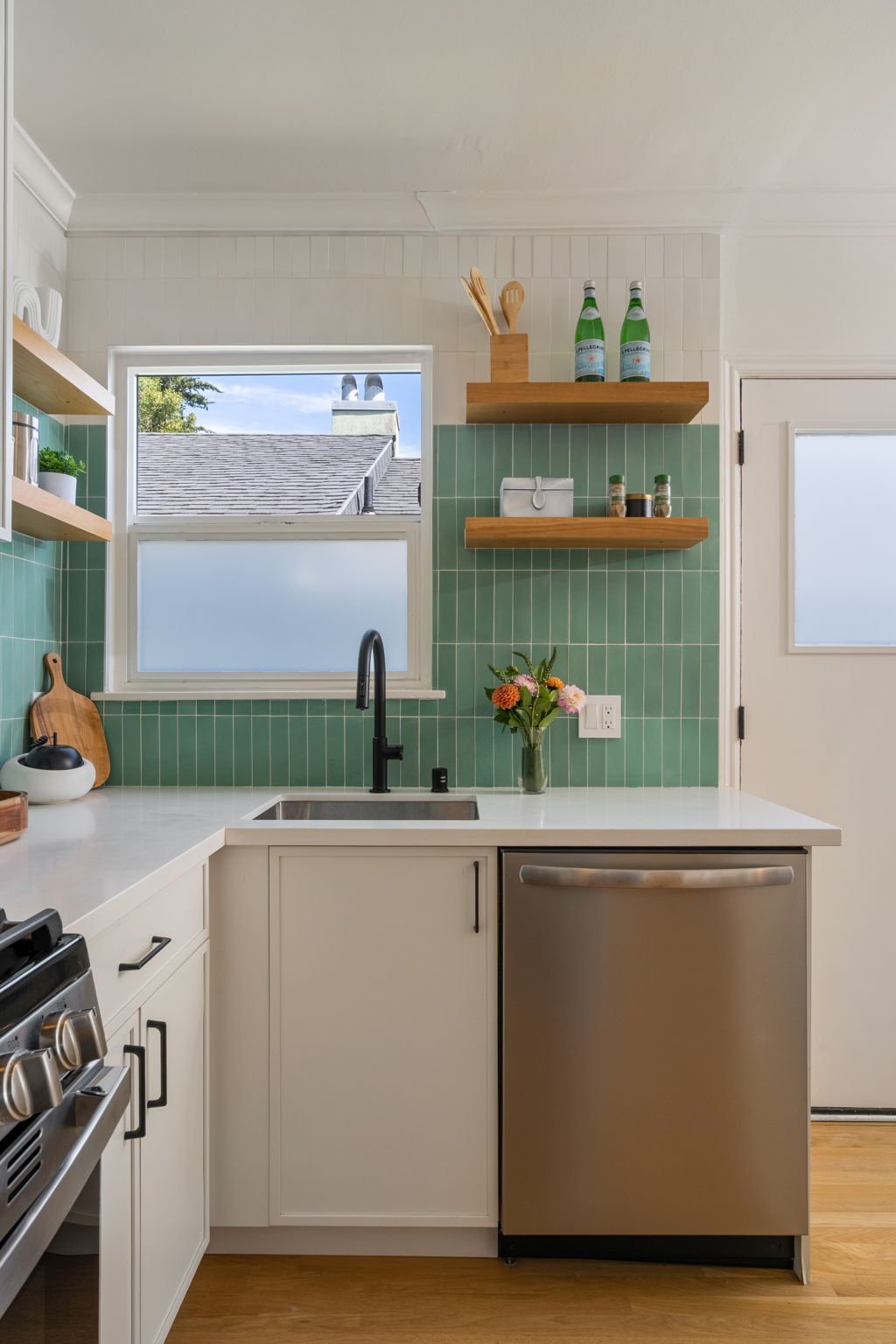 A modern kitchen with white cabinets, a stainless steel dishwasher, a black faucet, green tiled backsplash, a window showing a roof and blue sky, open wooden shelves with bottles and utensils, a vase with flowers, and a white door.