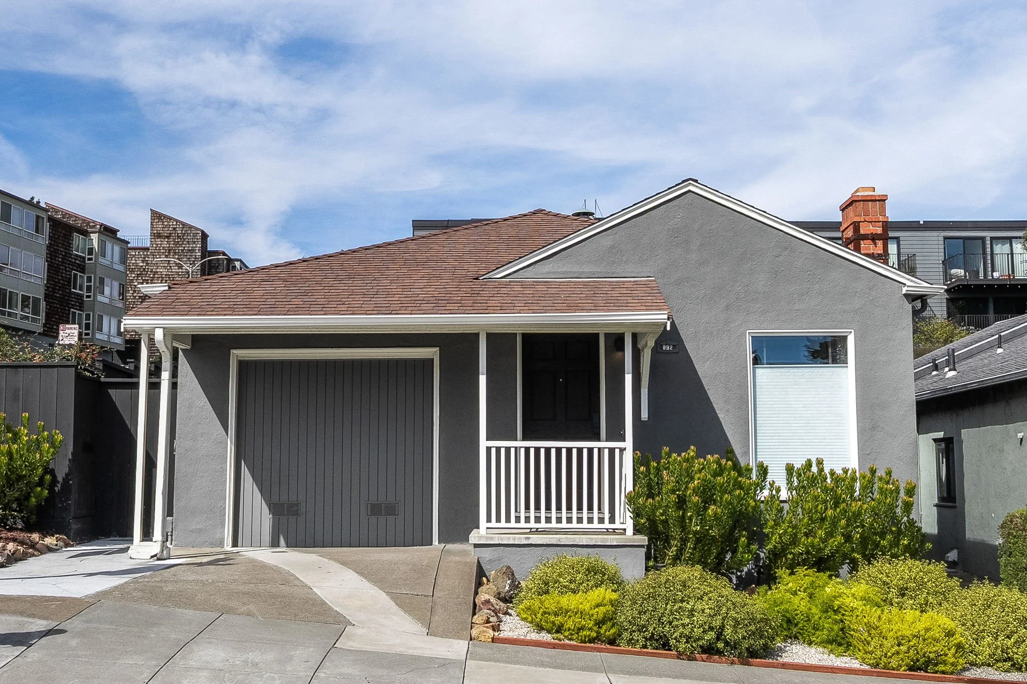 Front view of a modern gray house with a red shingle roof, a small porch, and landscaped plants in the front yard, with neighboring multi-story buildings and a partly cloudy sky in the background.