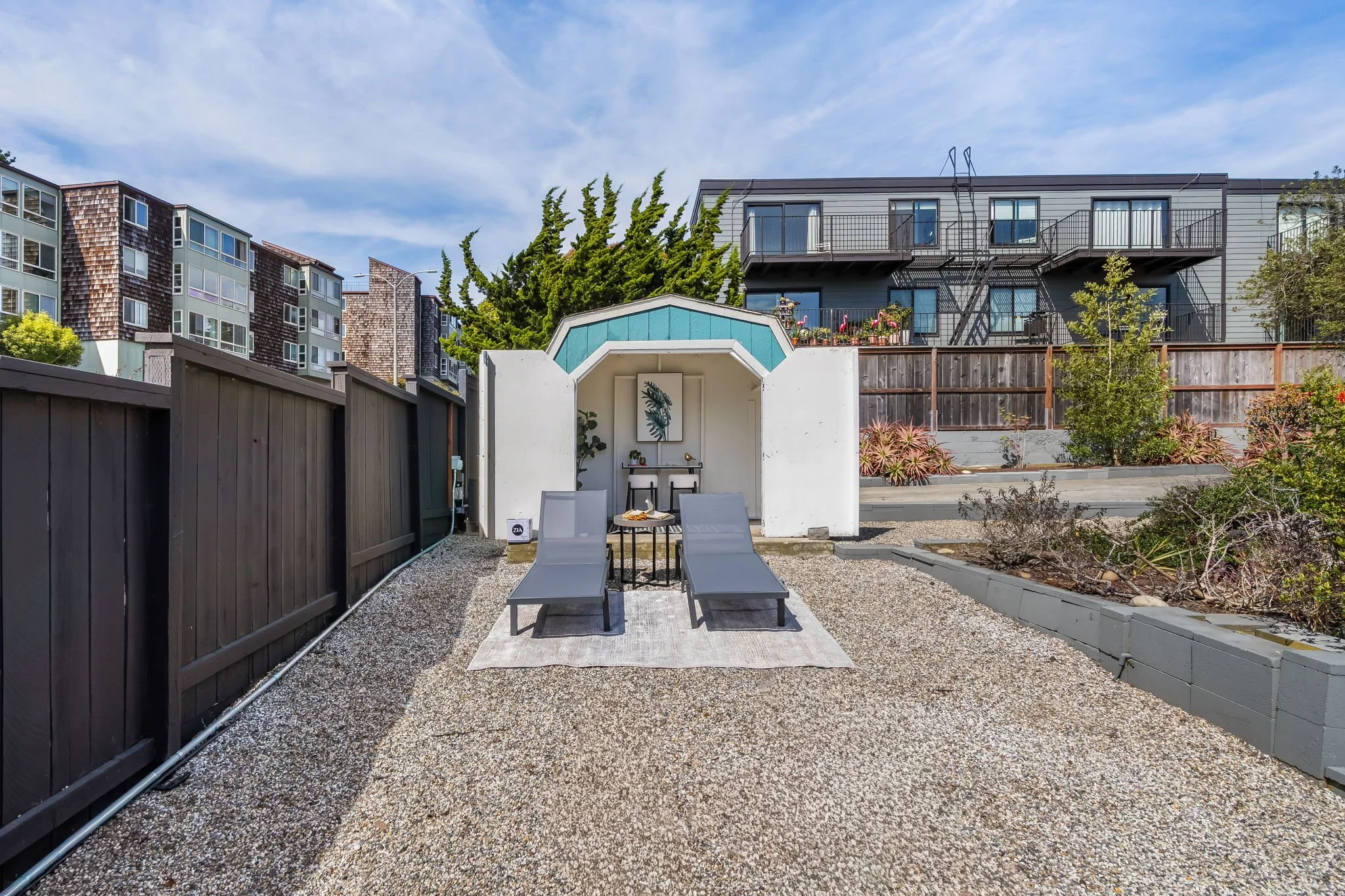 A backyard patio area with two lounge chairs, a small table, and a shelter with plants. Surrounding fencing and multi-story residential buildings in the background under a partly cloudy sky.