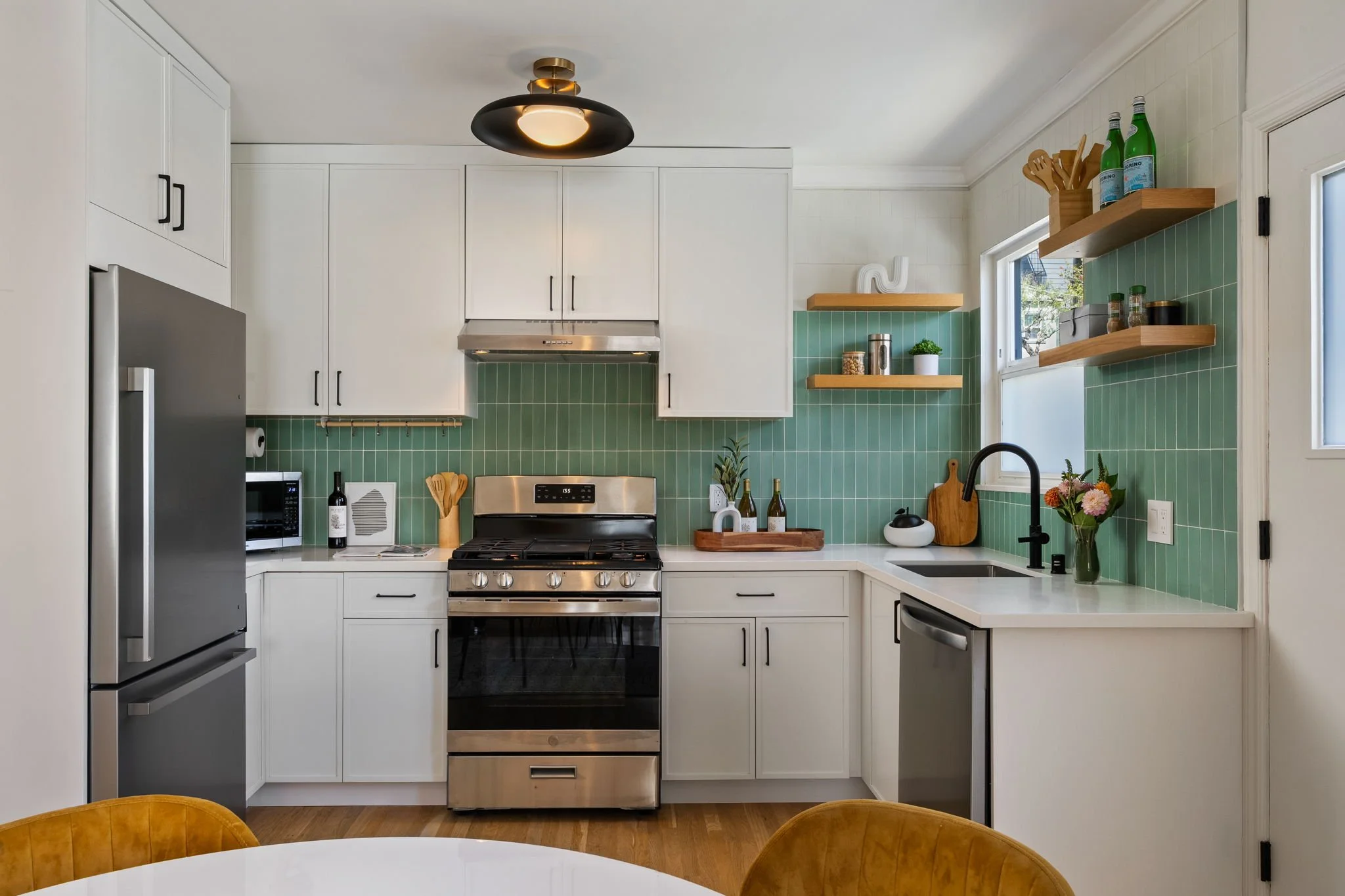 Modern kitchen with white cabinets, green tiled backsplash, stainless steel appliances, open wooden shelves, and a white countertop with a black sink. There are decorative items and plants on the shelves and countertop. A black ceiling light is also visible.