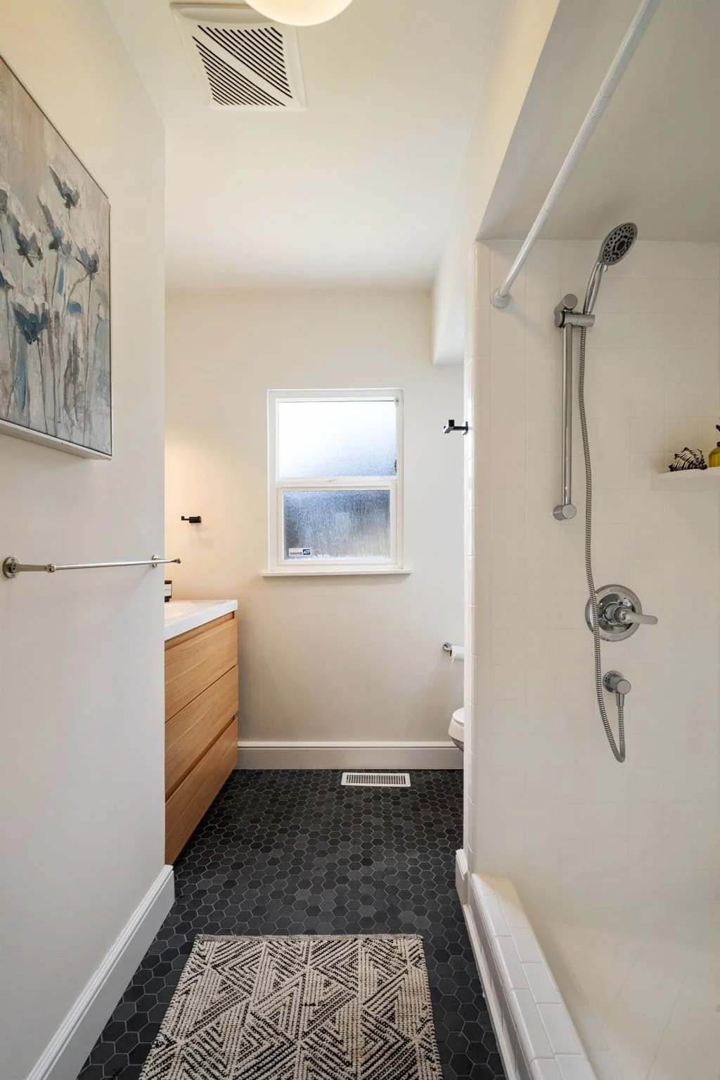 A small bathroom with a shower, window, and vanity with a wooden cabinet, featuring a black hexagon tile floor, a patterned rug, and artwork on the wall.