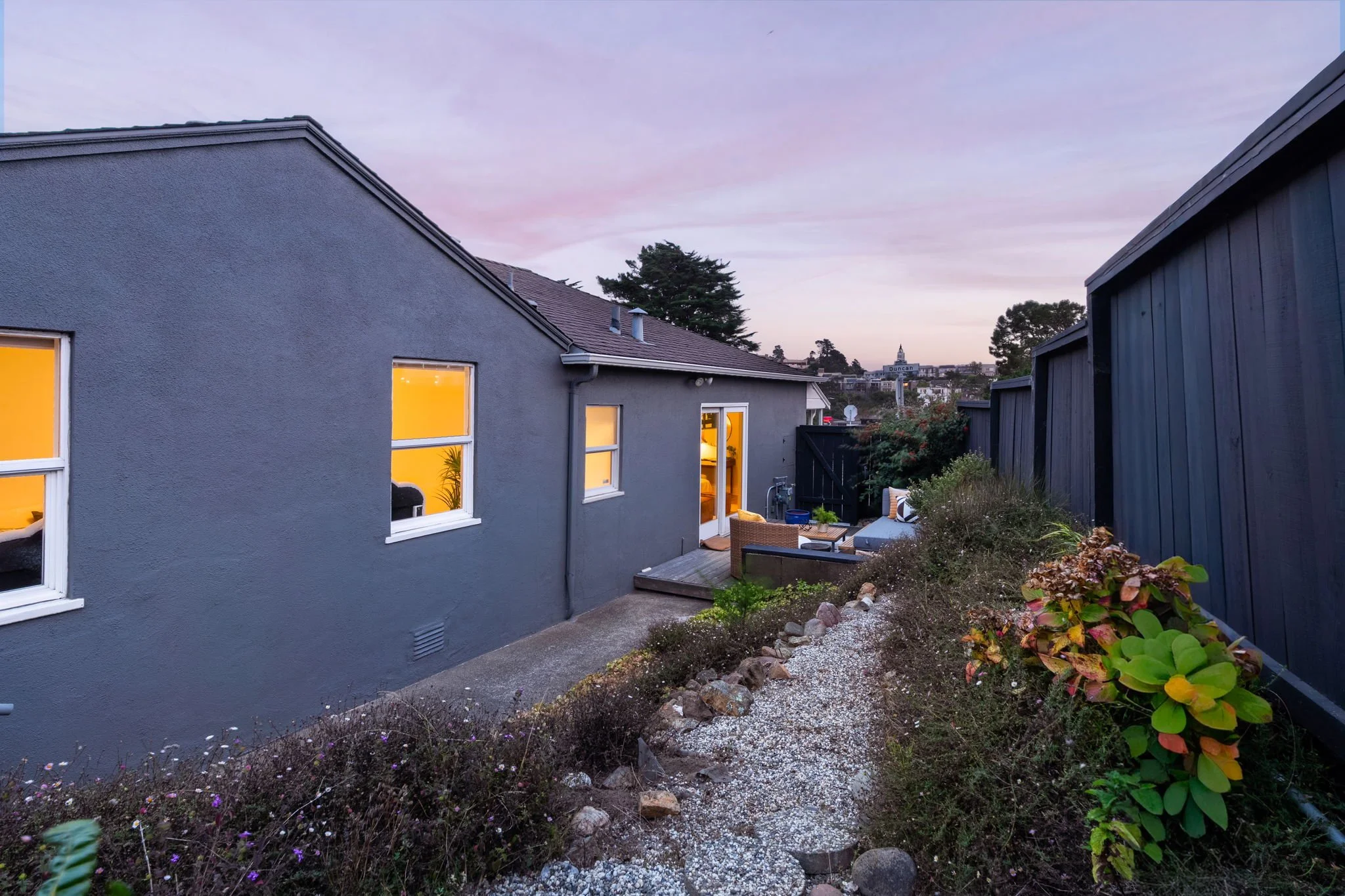 A backyard garden area with a gray house on the left, visible from the back, lit with warm yellow light from inside. The garden has a pathway with rocks and plants, and a seating area with outdoor furniture. A tall dark fence runs along the right sid