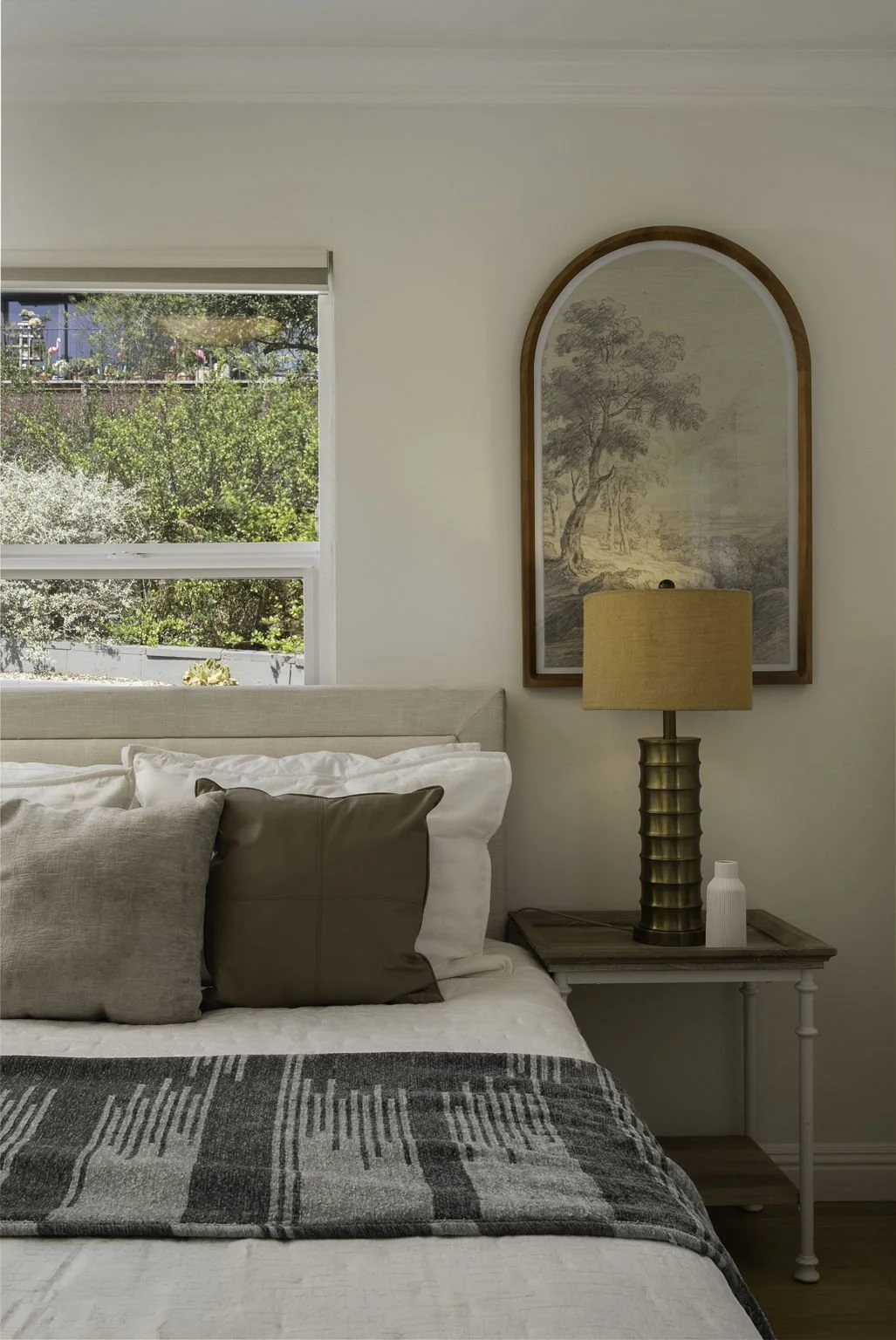 Close-up of a neatly made bed with multiple pillows, a bedside table with a textured lamp, a decorative vase, and framed artwork featuring a tree, with a window showing greenery outside.