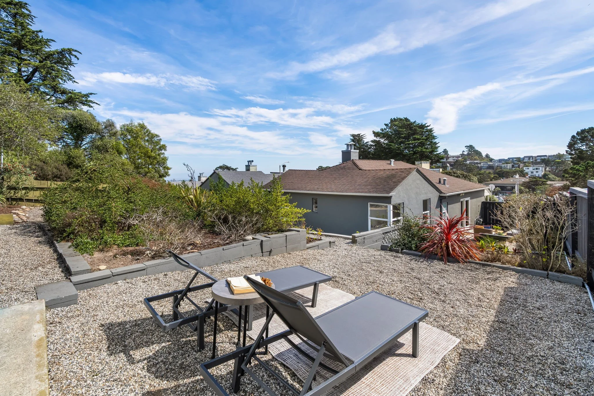 A backyard patio with two black lounge chairs and a small round table on a gravel surface, surrounded by plants and shrubs, with houses and trees in the background under a partly cloudy blue sky.