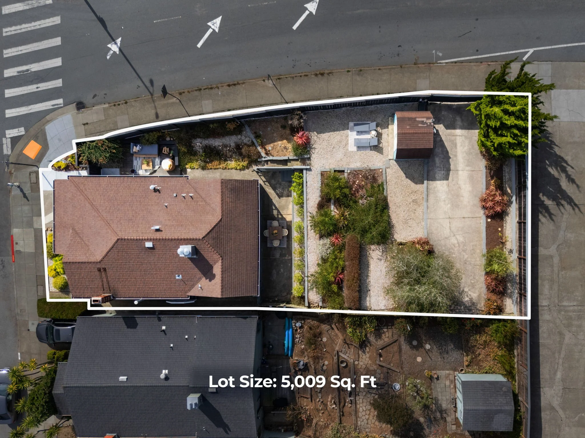 An aerial view of a residential lot with some plants, trees, and a small shed, surrounded by houses, streets, and a sidewalk. The lot size is 5,009 square feet.