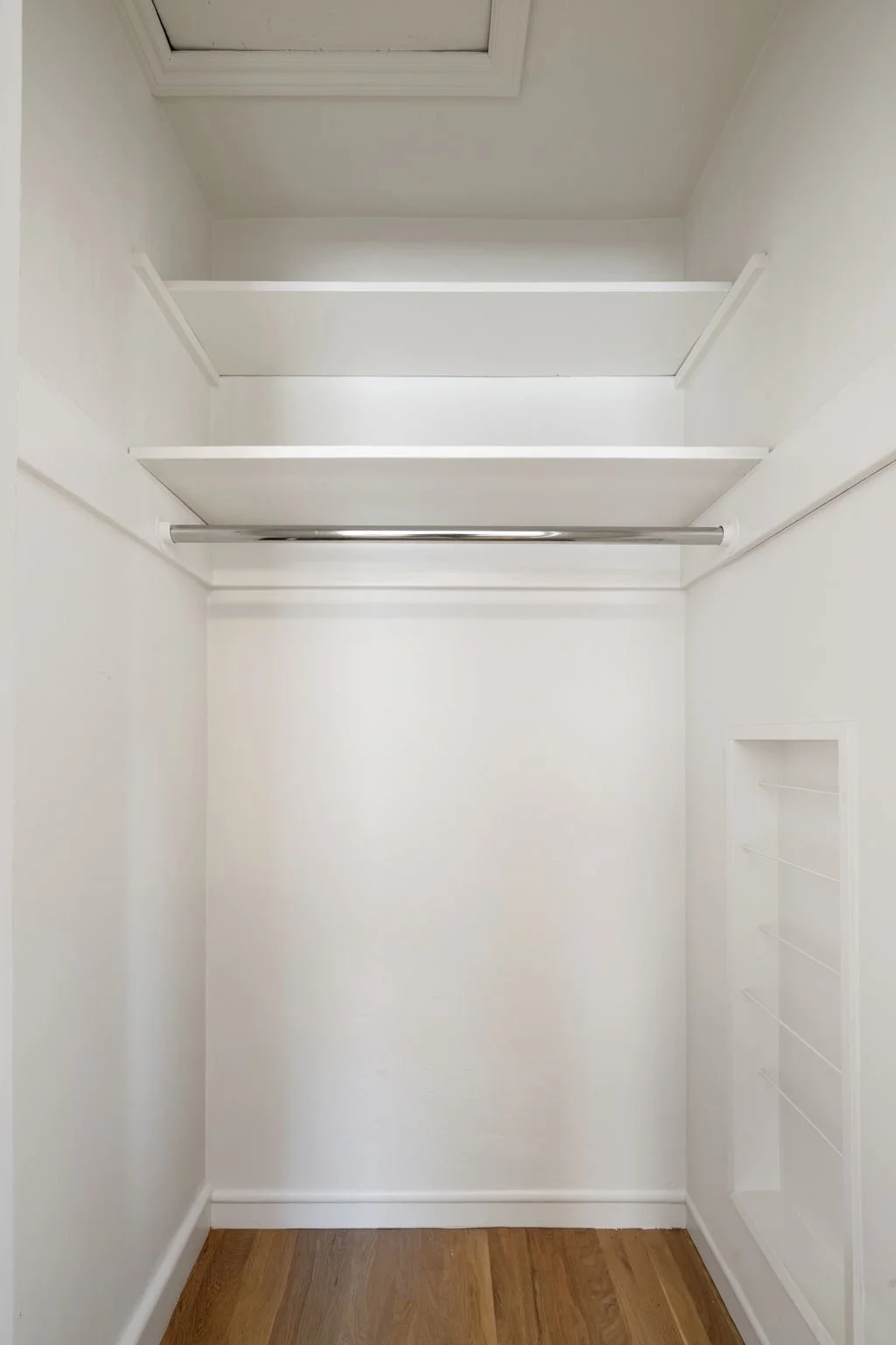 Empty walk-in closet with white walls, shelves, a hanging rod, and a small vent or niche on the right wall, with wooden floor.