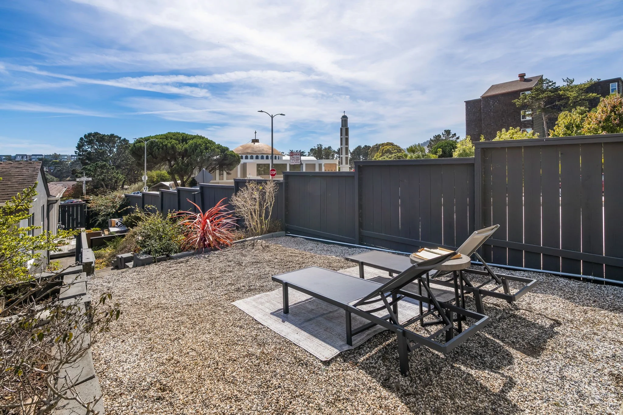 A backyard patio area with two lounge chairs on a rug, surrounded by gravel and plants, with a wooden fence, trees, a church with a steeple, and a blue sky with clouds in the background.