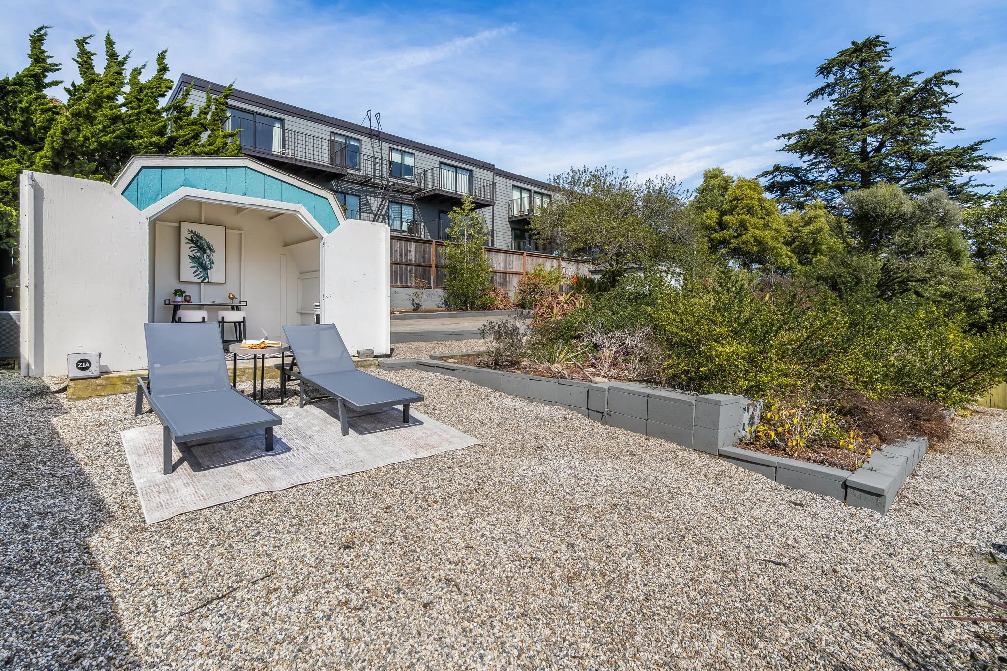 A backyard patio with two lounge chairs, a small table, and a white and blue decorative structure. In the background, there is a modern multi-story apartment building with balconies and a variety of trees and shrubs.