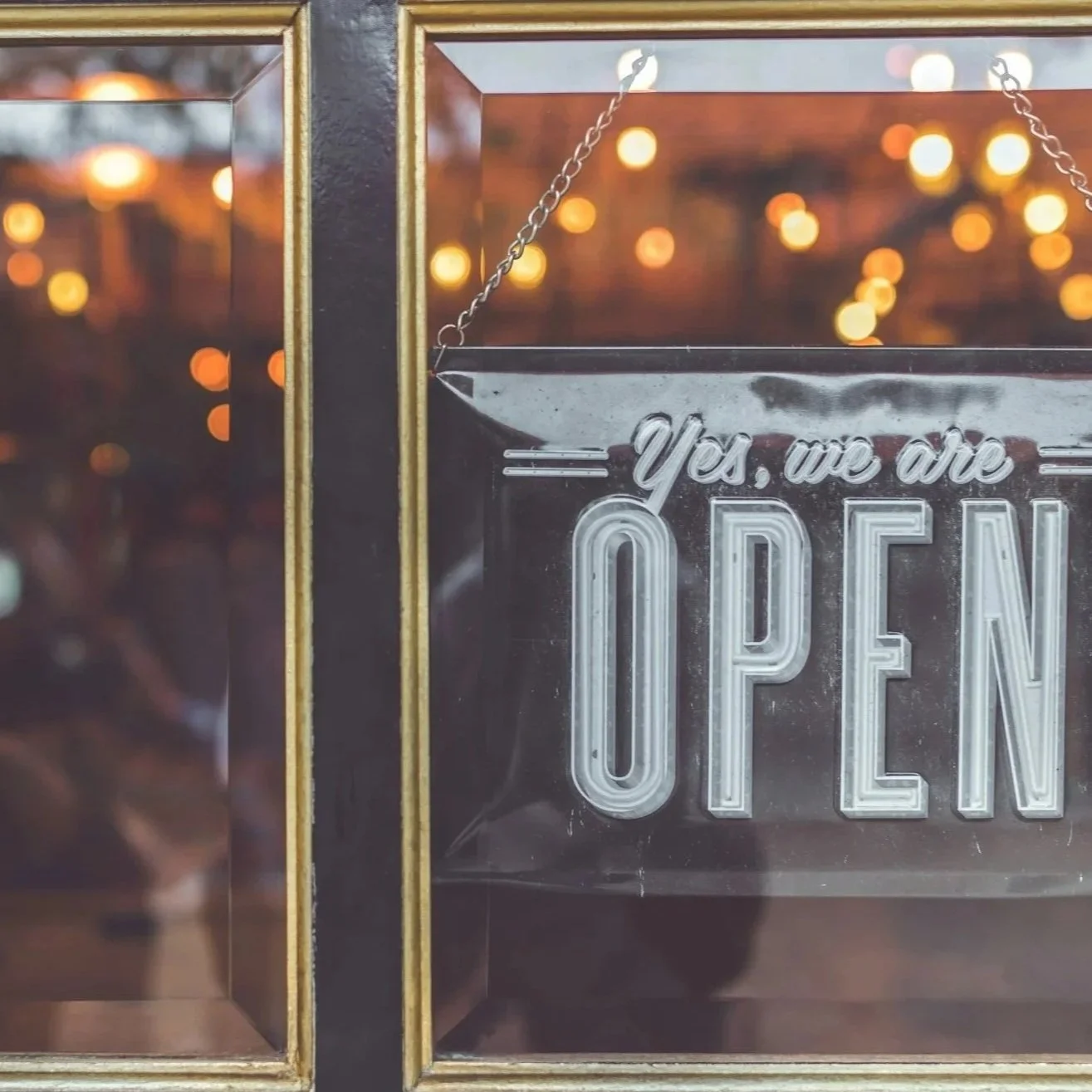 Close-up of a vintage framed sign reading “Yes, we are OPEN,” with warm string-light reflections.