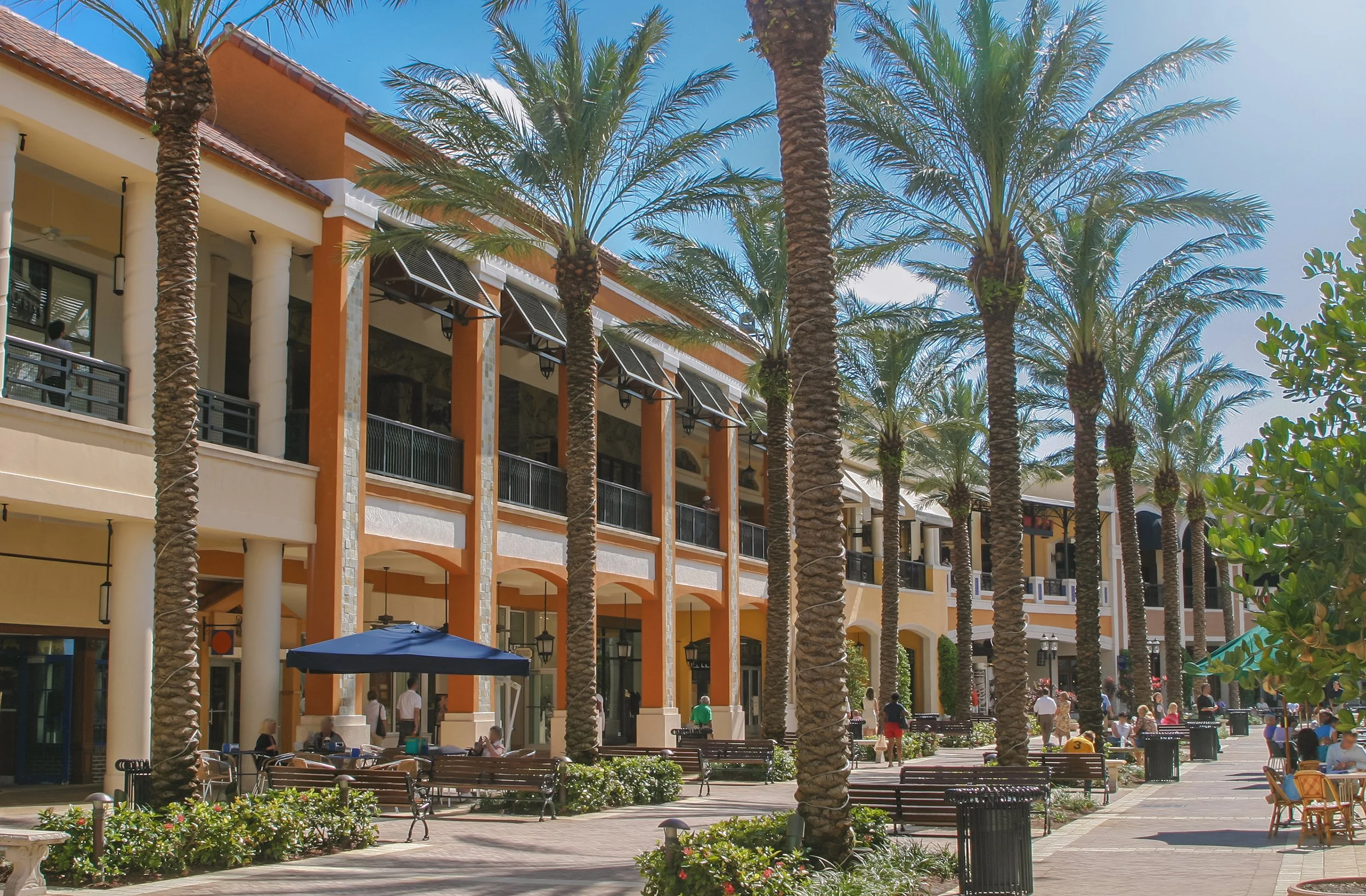 Outdoor shopping or dining plaza with palm trees, benches, umbrellas, and stores. People walking and sitting in the area under a bright blue sky.