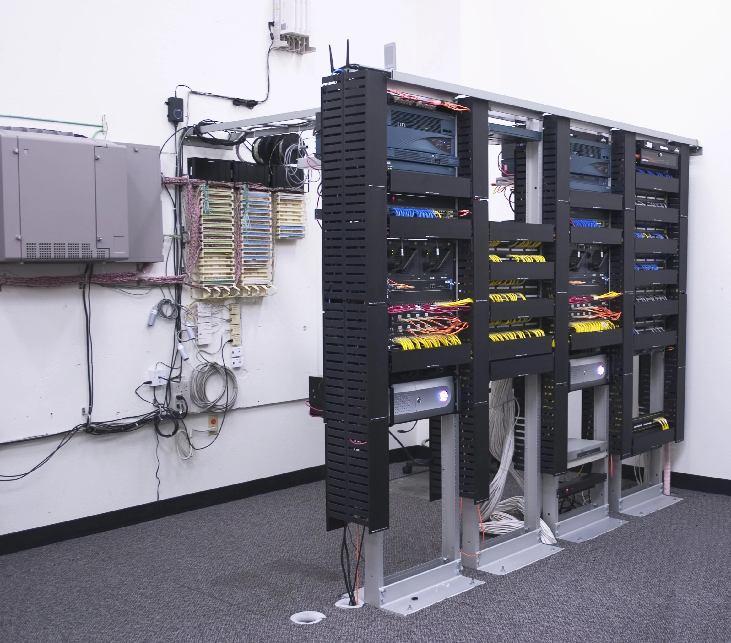 Server racks with network cables and equipment mounted against a white wall, with a gray electrical box and organized color-coded cable bundles nearby.