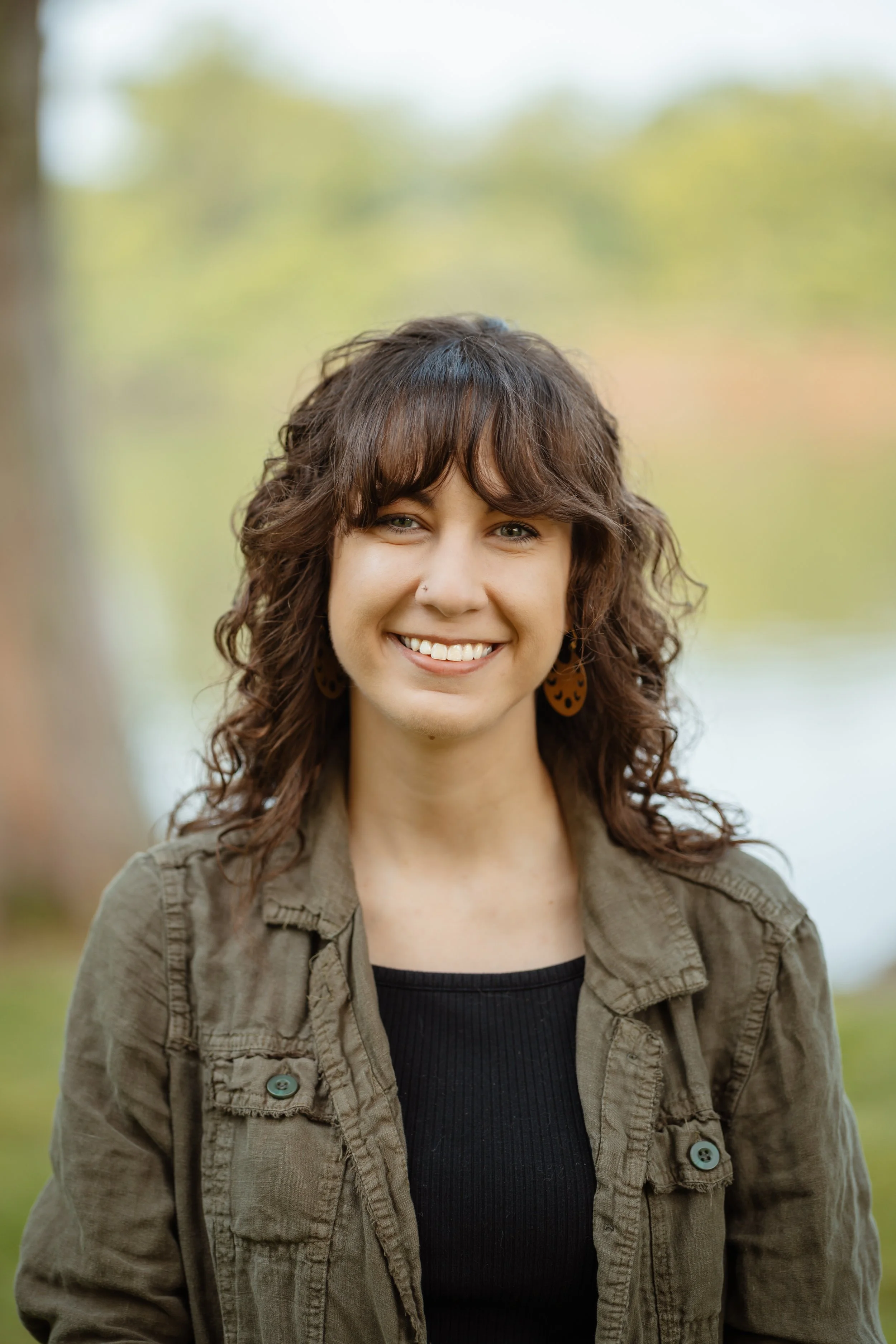 A close-up portrait of a woman with curly brown hair, green eyes, and a smile, standing outdoors with a blurred background of trees and sky.