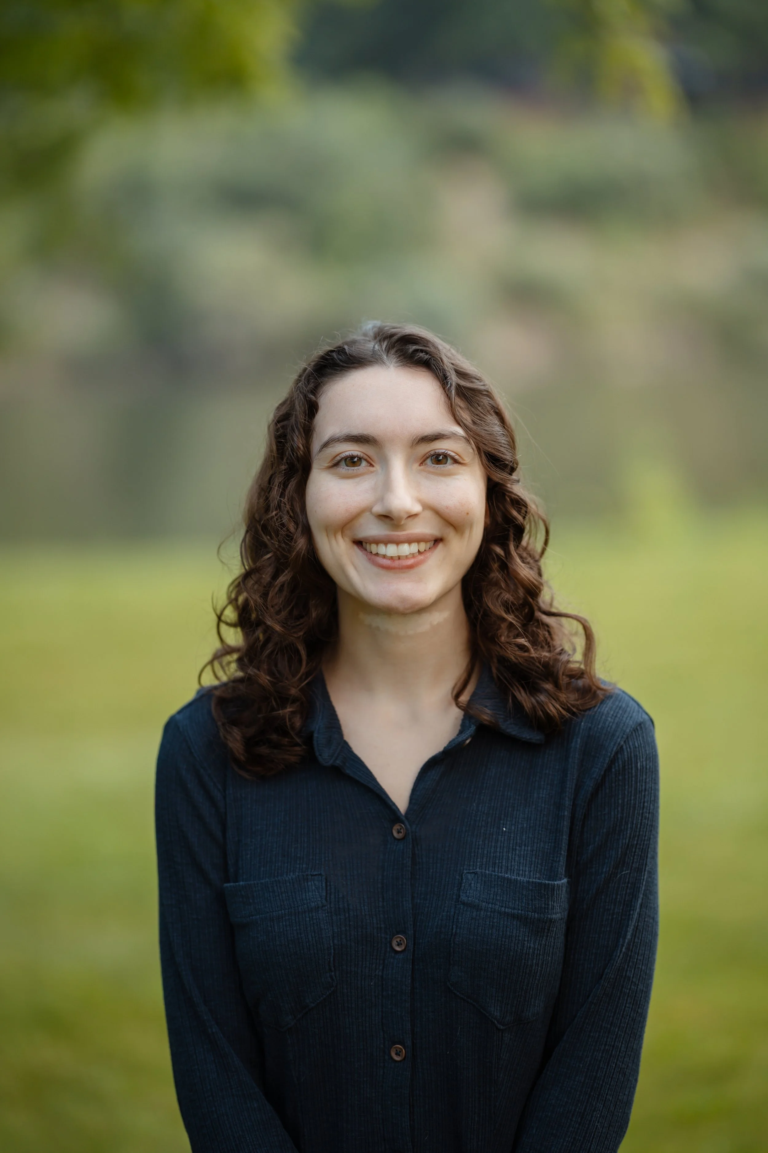 A young woman with curly brown hair and a wide smile wearing a dark blue shirt outdoors with a blurred green and yellow background.