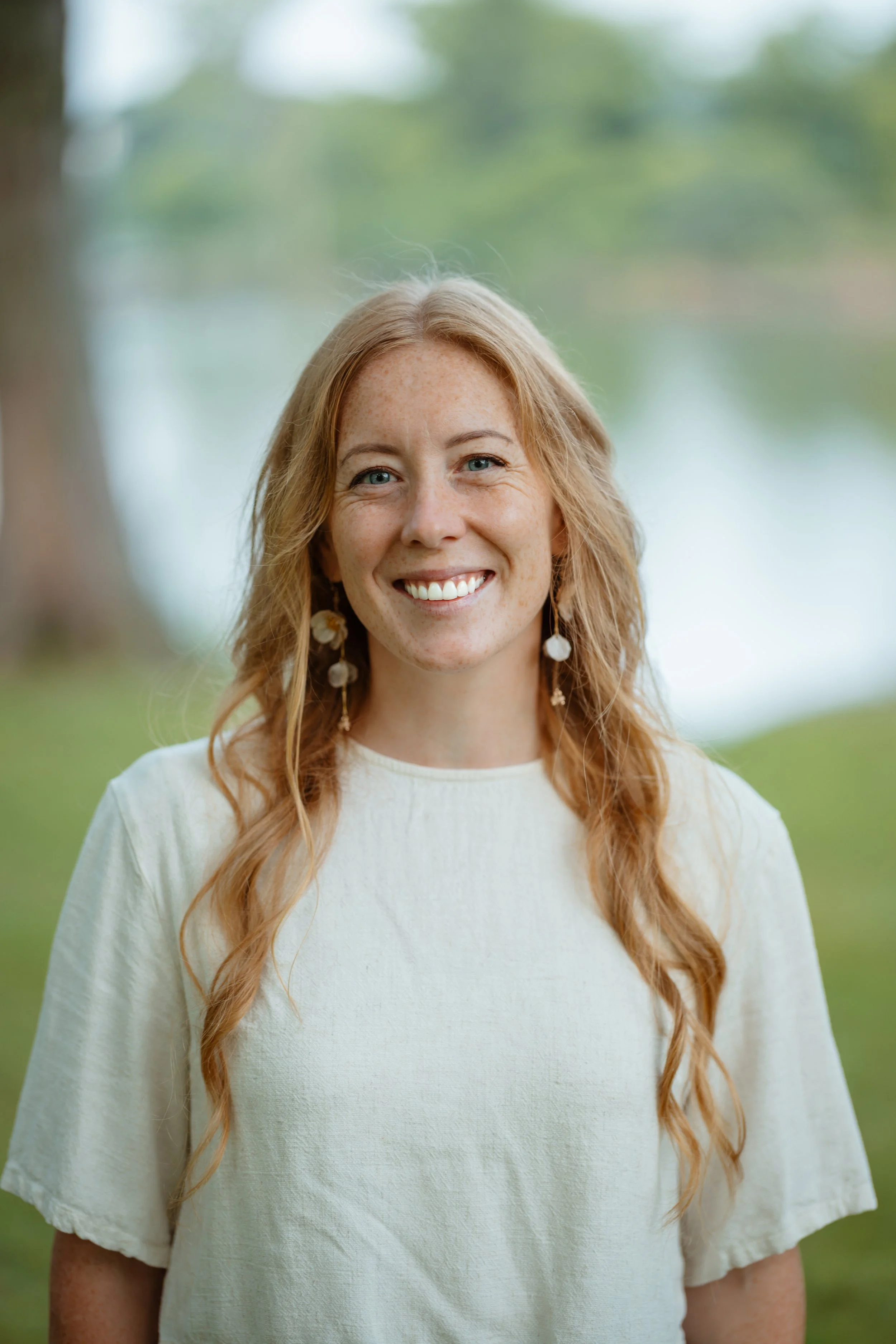 Close-up of a woman with long, wavy red hair, smiling, wearing earrings and a cream-colored blouse, outdoors with blurred green trees in the background.