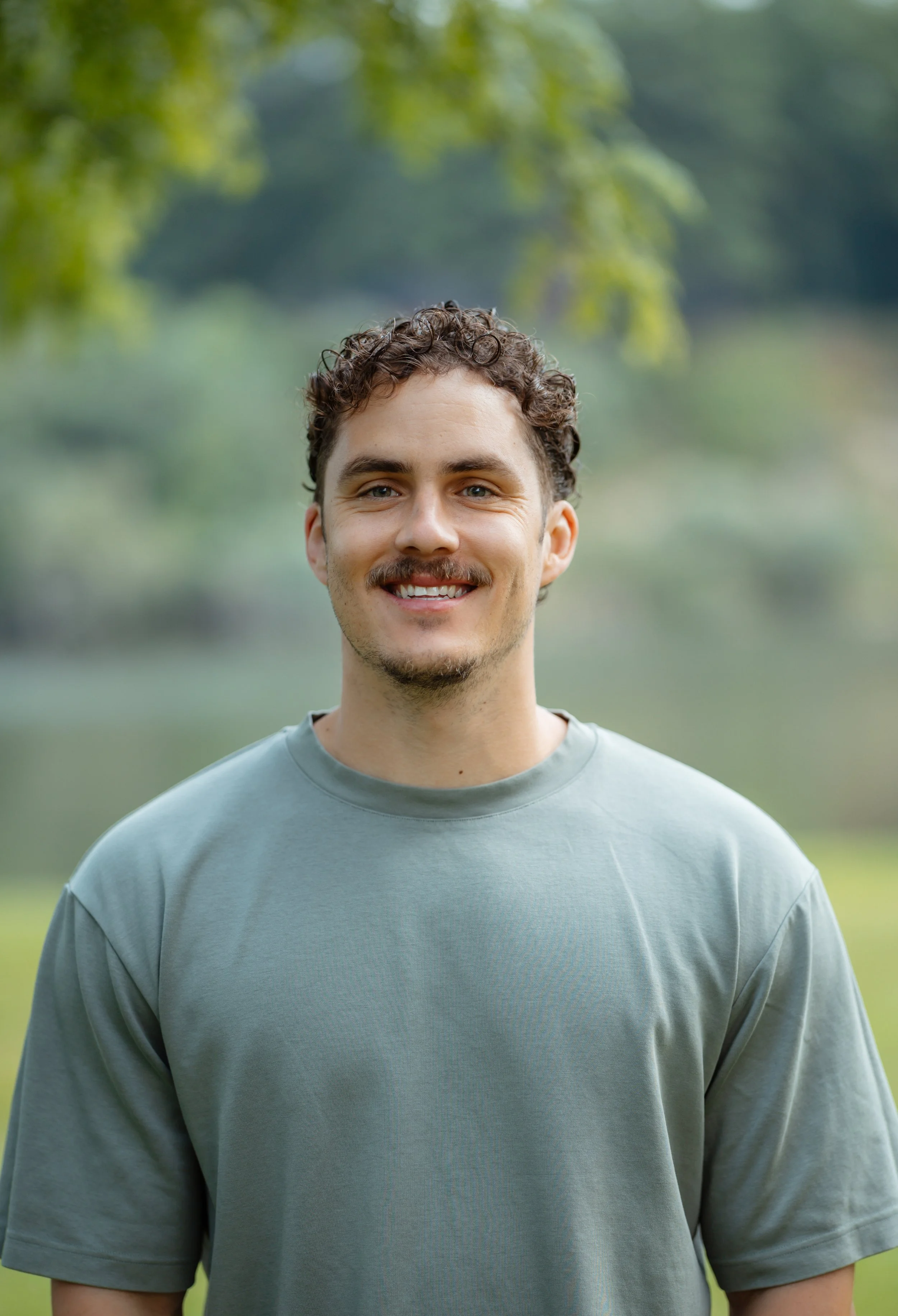A smiling young man with curly brown hair and light skin, wearing a gray sweatshirt, standing outdoors near a body of water with green trees in the background.