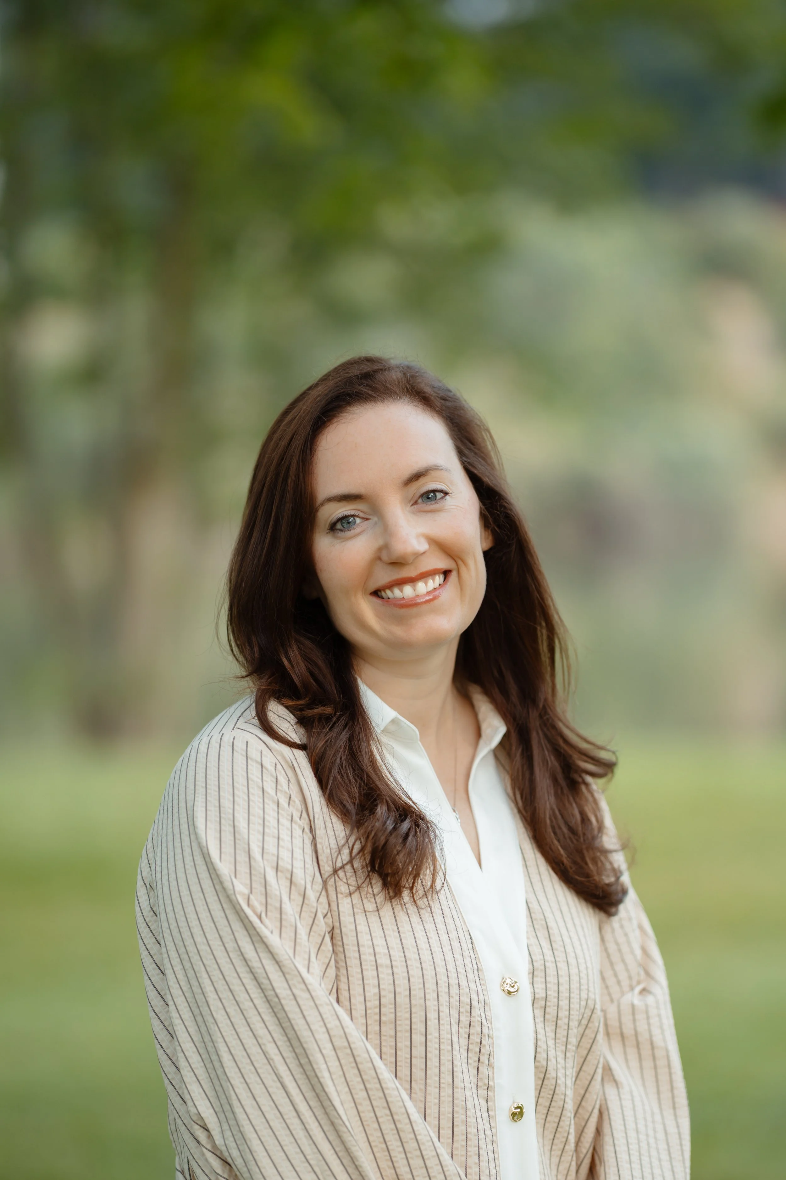 A woman with long brunette hair, light skin, and blue eyes smiling outdoors with a background of green trees and blurred foliage.
