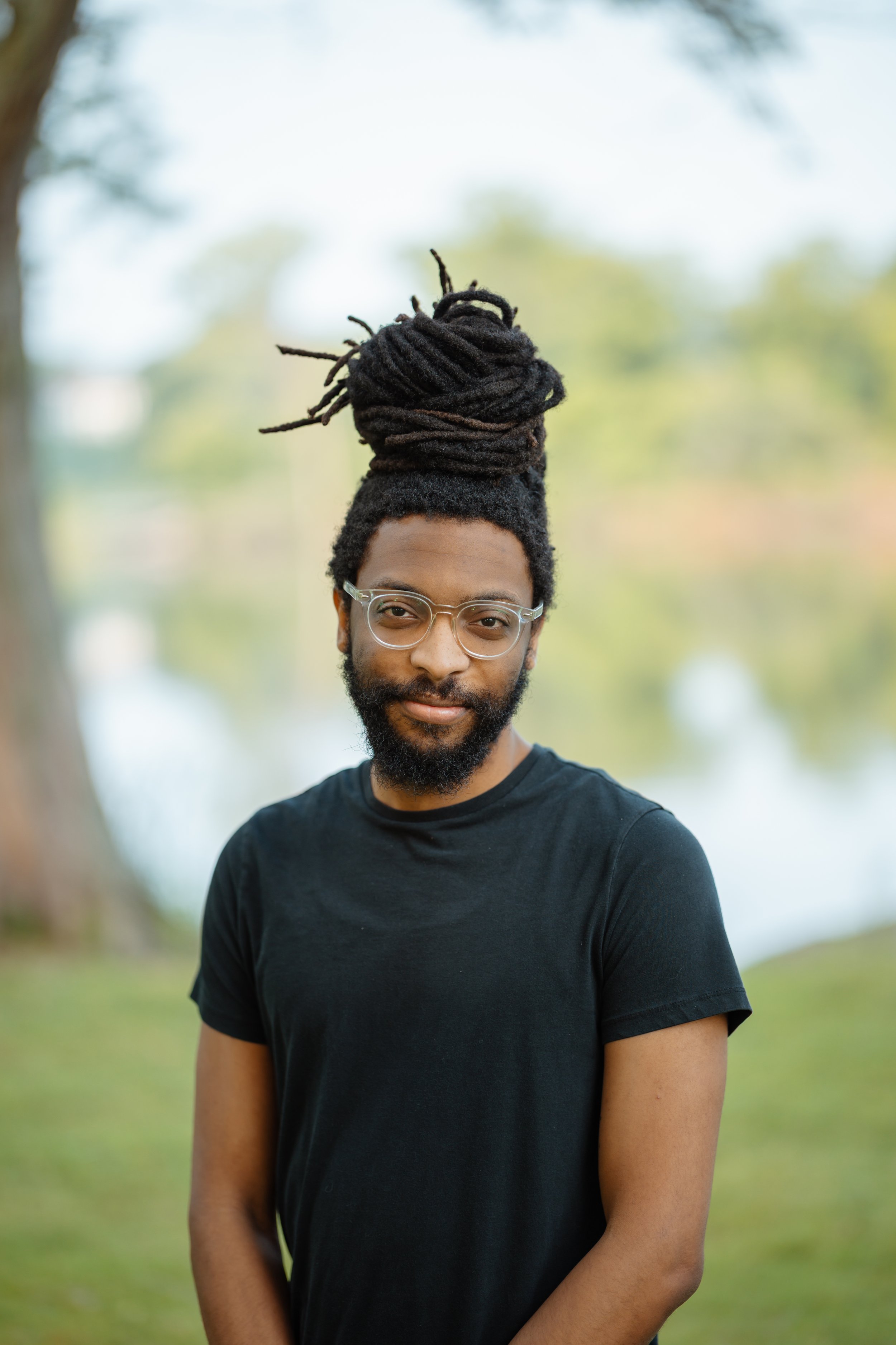 A man with black dreadlocks tied in a bun, glasses, beard, wearing a black t-shirt outdoors with trees and water in the background.
