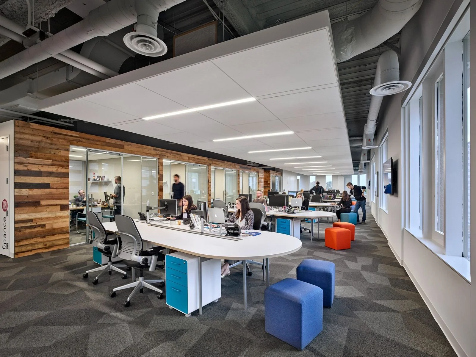 Open-plan modern office with employees working at desks, some in glass-walled meeting rooms, featuring a wooden accent wall, large windows, and colorful seating.