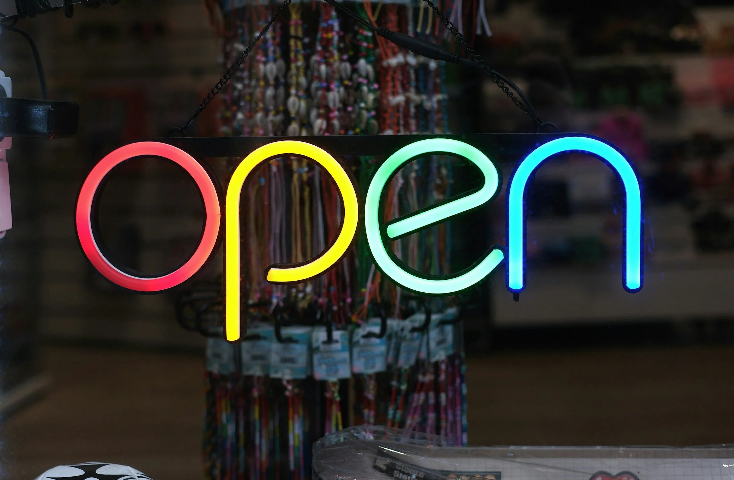 Neon sign displaying the word 'open' in red, yellow, green, and blue colors, hanging in front of racks of jewelry or accessories in a store.