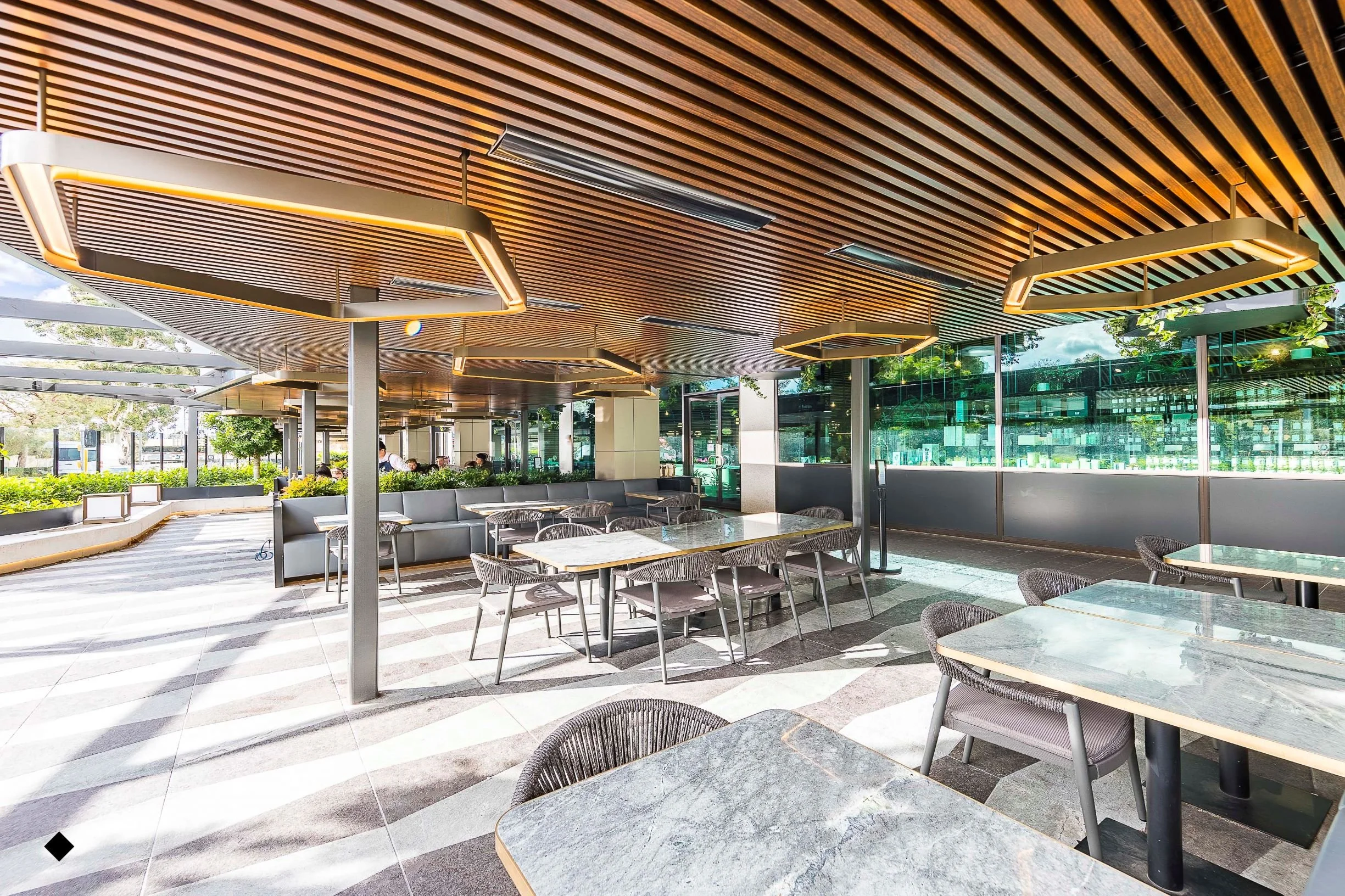 Outdoor seating area at a modern cafe with wooden ceiling panels, square ceiling lights, glass walls, and trees outside.
