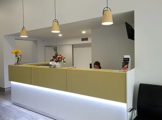 Reception desk with yellow panels, white base with LED lighting, three hanging pendant lights, a vase with flowers, and brochures, with a person partially visible behind the desk and a wall-mounted TV to the right.