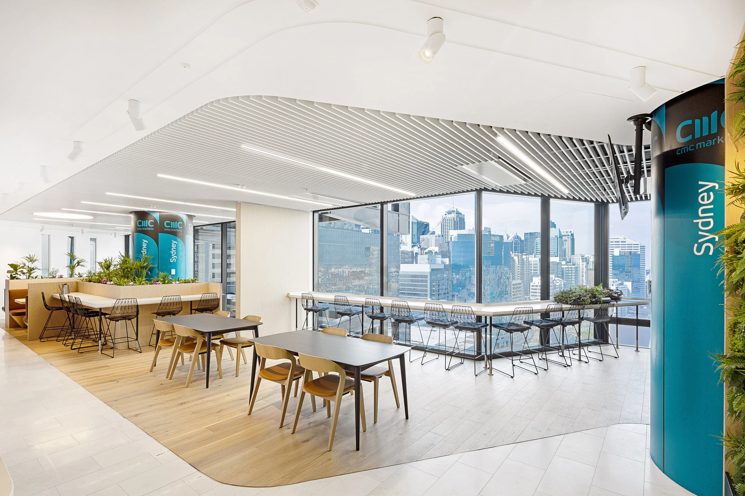 Modern office cafeteria with large windows and city view, tables and chairs, indoor plants, ceiling with white slats and spotlights.