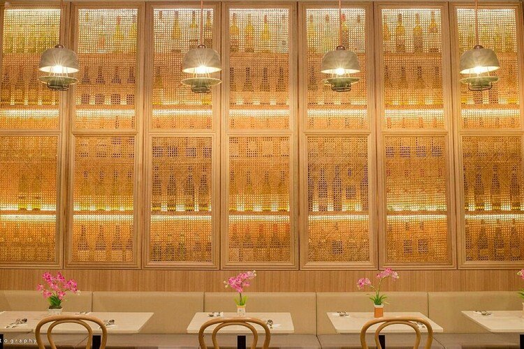 Indoor dining area with a long beige bench, three white tables, and six wooden chairs. Each table has a small potted pink orchid. The wall behind features a large decorative wooden panel with warm lighting and three hanging lamps.