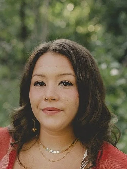 A woman with shoulder-length brown hair, wearing a red top, gold earrings, and layered necklaces, standing outdoors with green trees in the background.