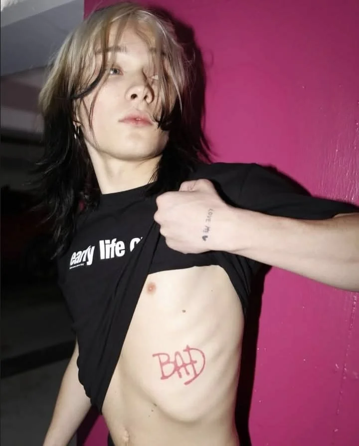 A young person with long, wavy hair lifts their black T-shirt to reveal a red tattoo on their side that says "BAD" with a circle around it. They are standing against a pink wall, with some hair covering part of their face.