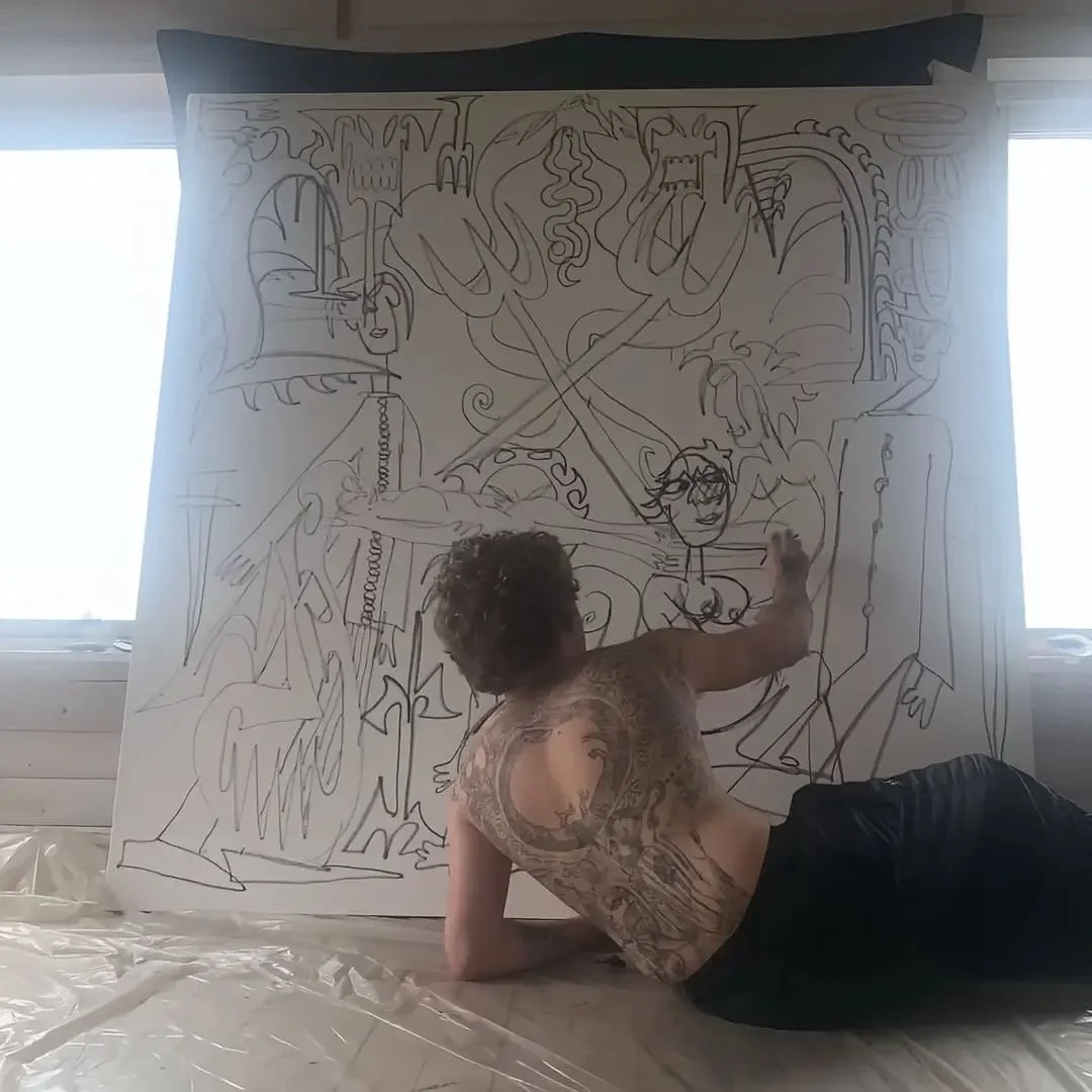 A person with tattoos on their back, sitting on the floor and drawing a large abstract mural on a white canvas. The mural includes various abstract shapes, faces, and figures, with windows visible on either side of the canvas.