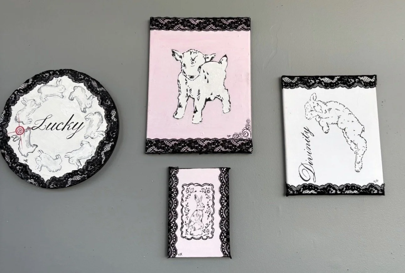 Four framed drawings of rabbits, some with words like "Lucky" and "Dainty," hanging on a gray wall, decorated with lace borders.