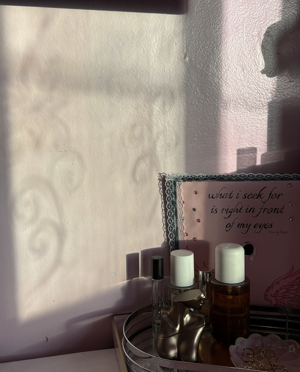 A pink framed quote on a pink wall that says, "what i seek for is right in front of my eyes" by Young Lean. In front of the frame, there are bottles including perfume and skincare, a small metallic mannequin bust, and jewelry on a circular tray.