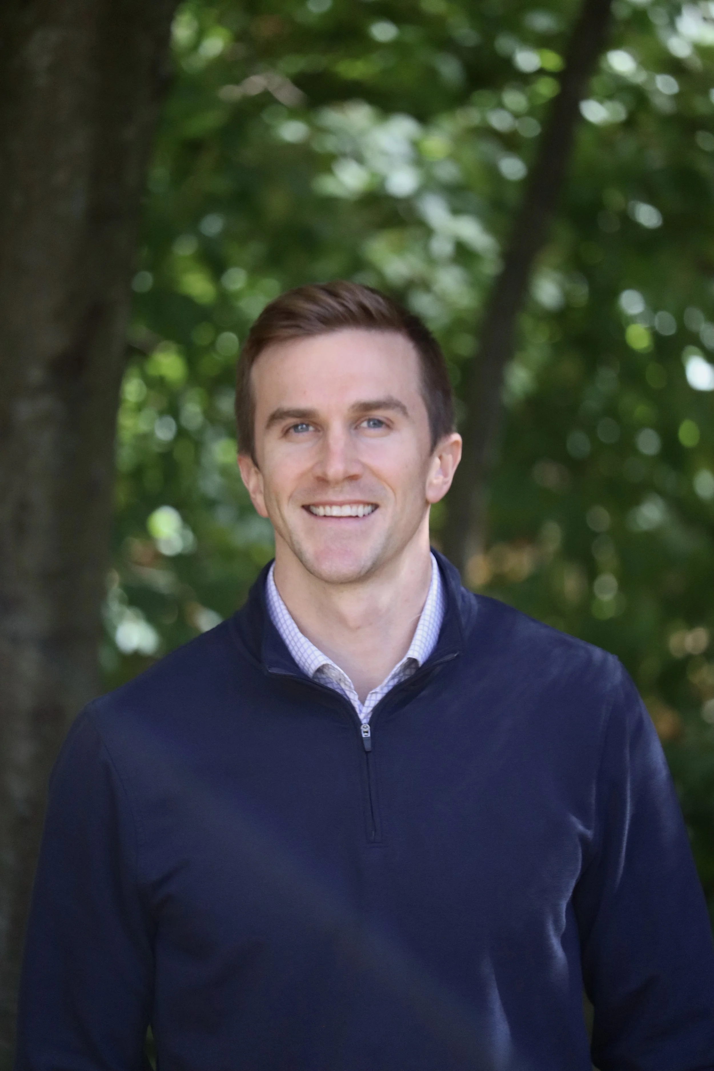 A smiling man with short brown hair and blue eyes, wearing a navy sweater over a collared shirt, standing outdoors with blurred green foliage in the background.