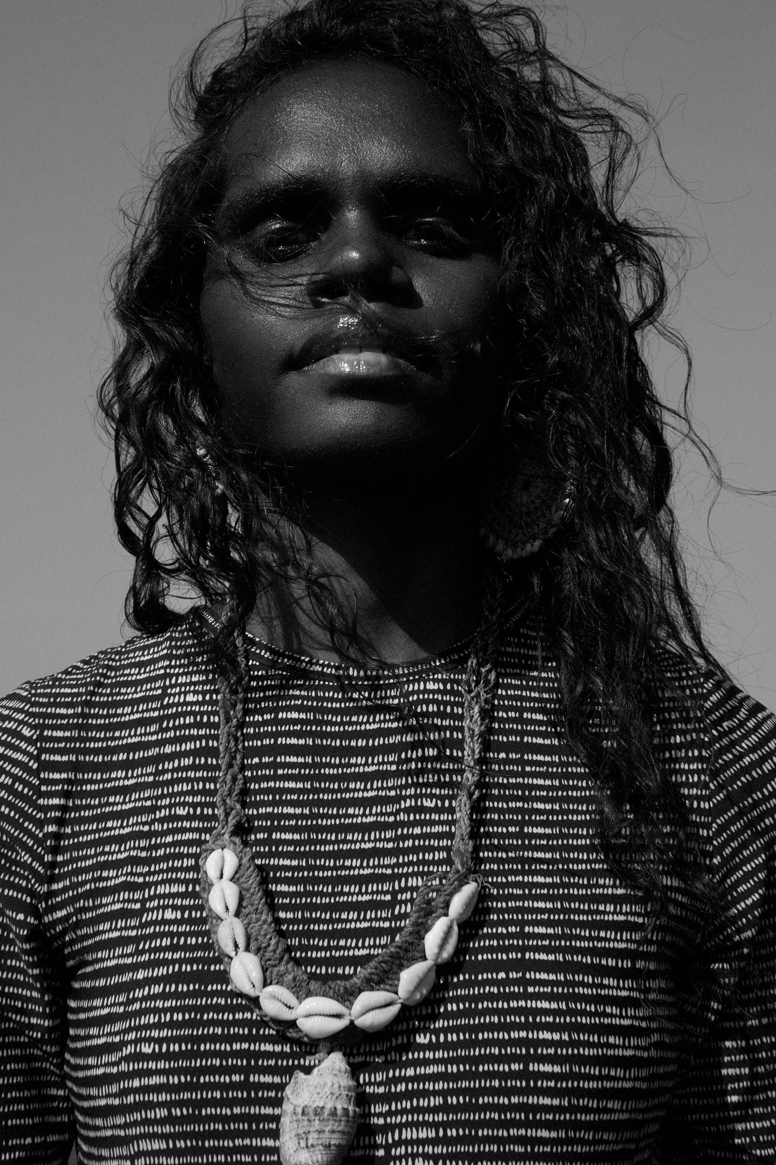 A woman with long curly hair, wearing a patterned top and shell necklaces, looking directly at the camera with a serious expression in black and white.