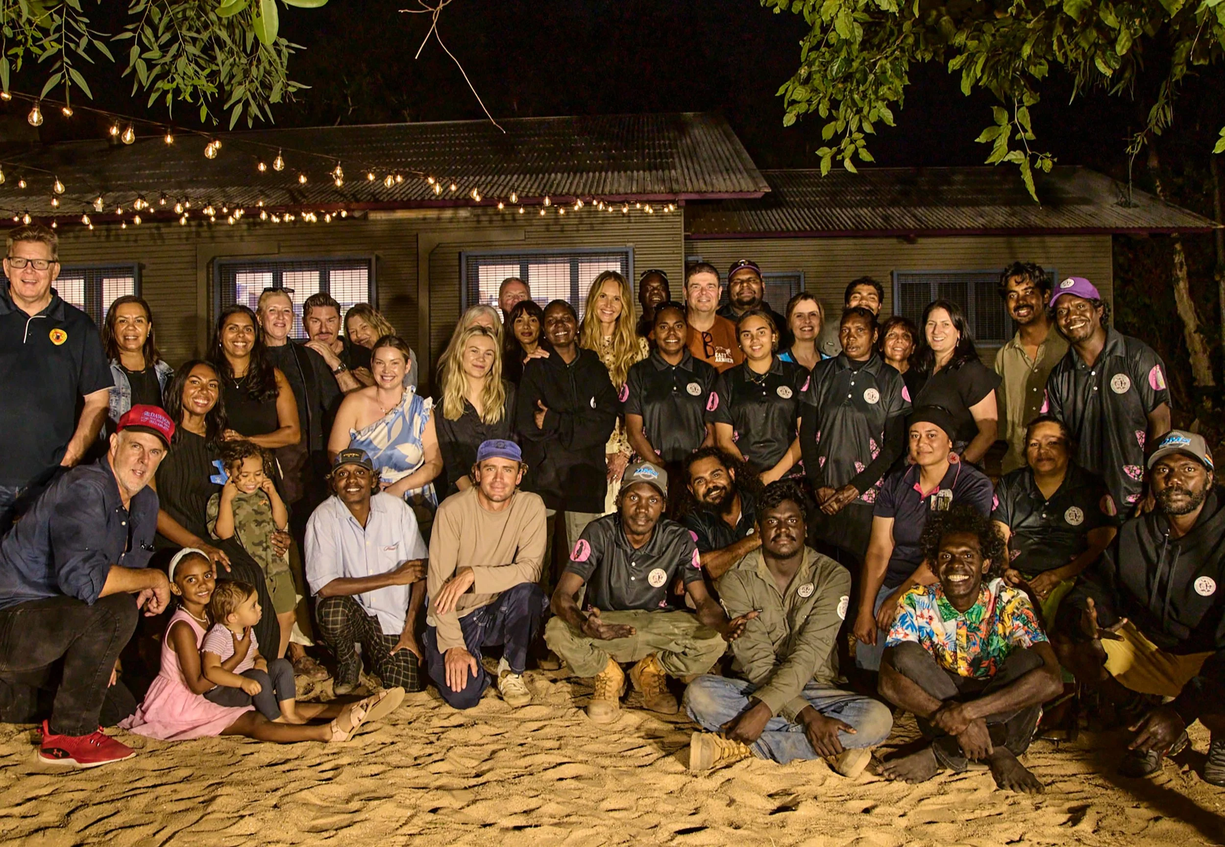Group of diverse people posing for a photo outdoors at night, with string lights hanging overhead and a wooden building in the background.