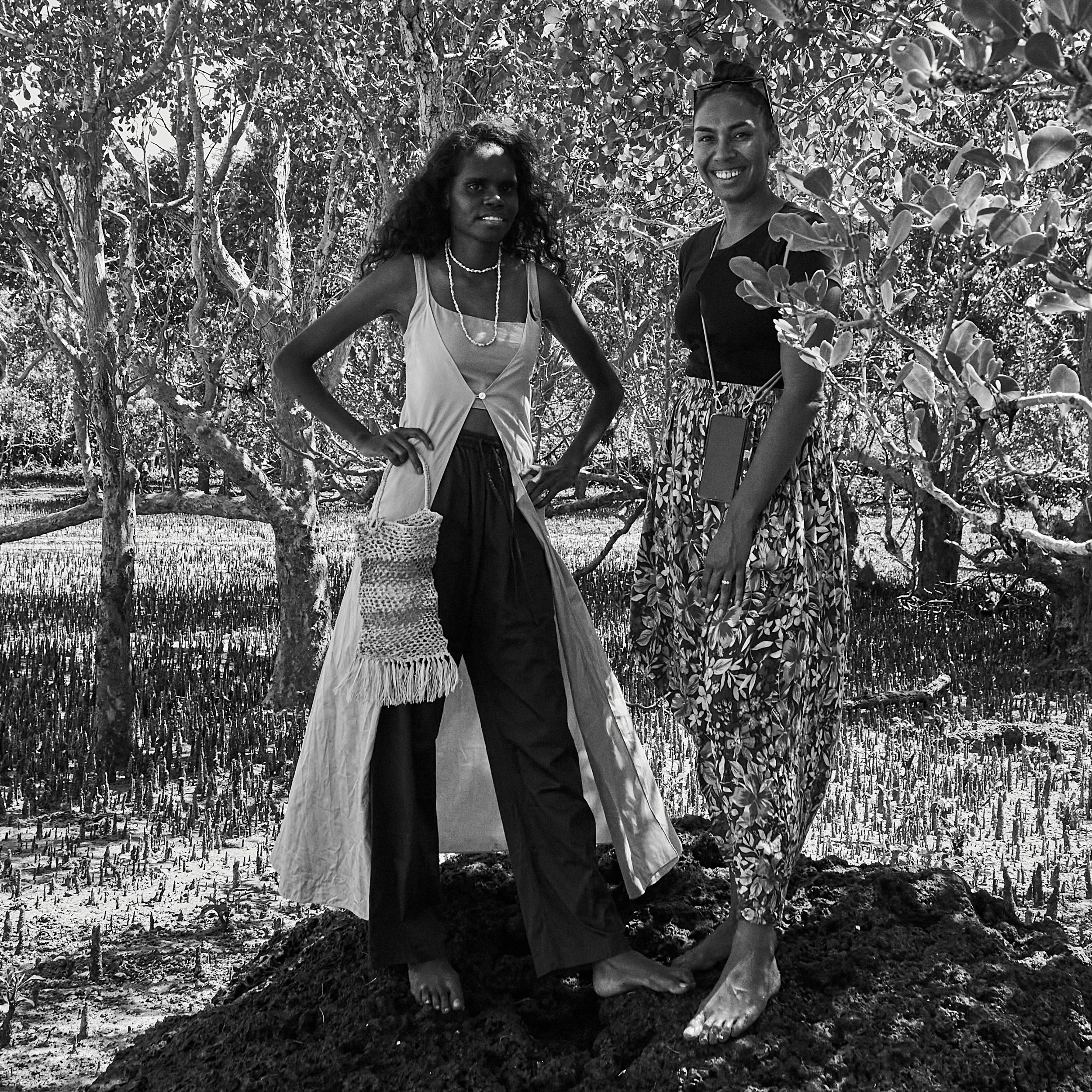 Black-and-white photograph of Tarlisa Gaykamangu and Liandra Gaykamangu standing outdoors among trees, one wearing a sculptural costume and the other smiling beside her.