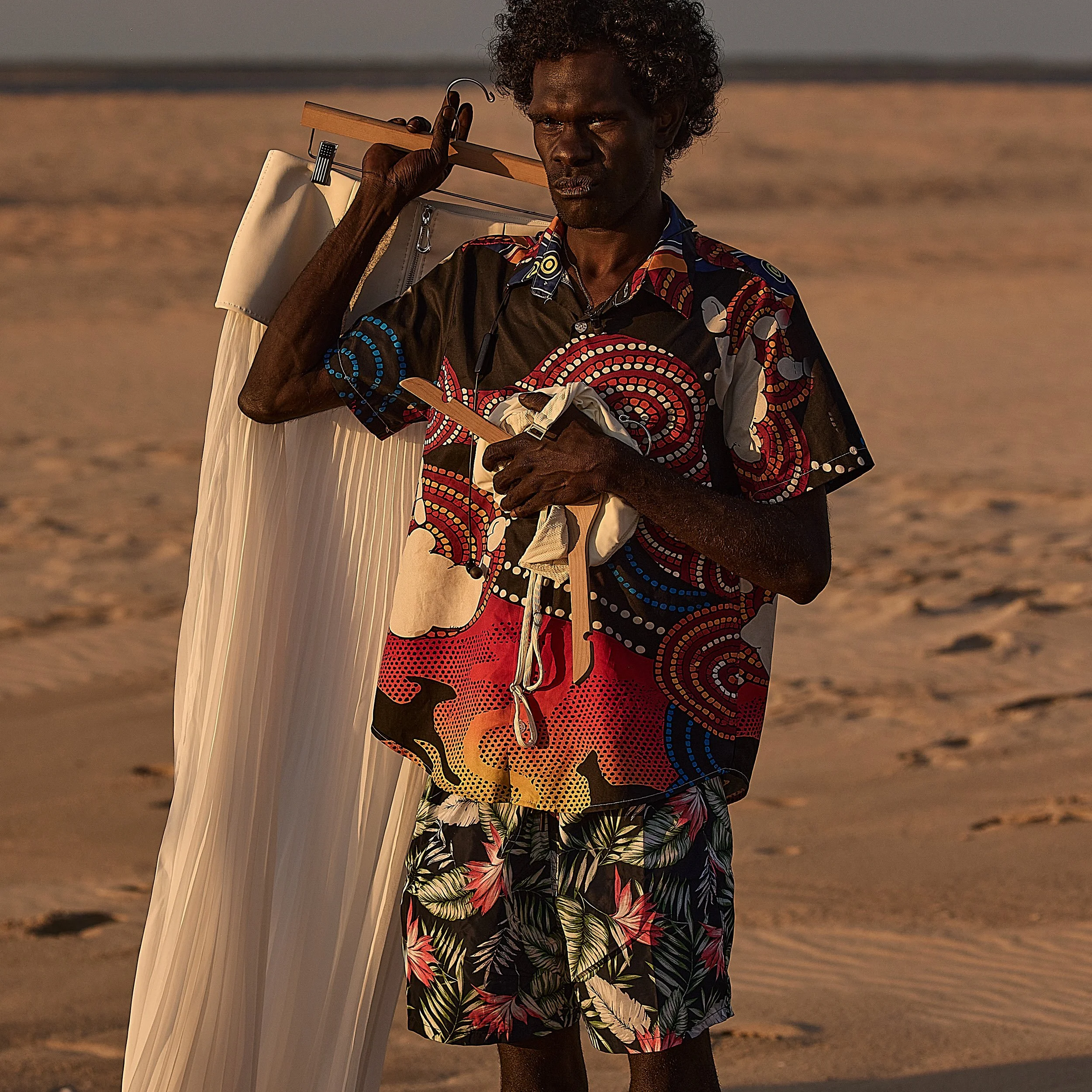 Jack Minmidhi walking along a beach in East Arnhem Land, carrying a garment bag and wooden hanger over his shoulder.