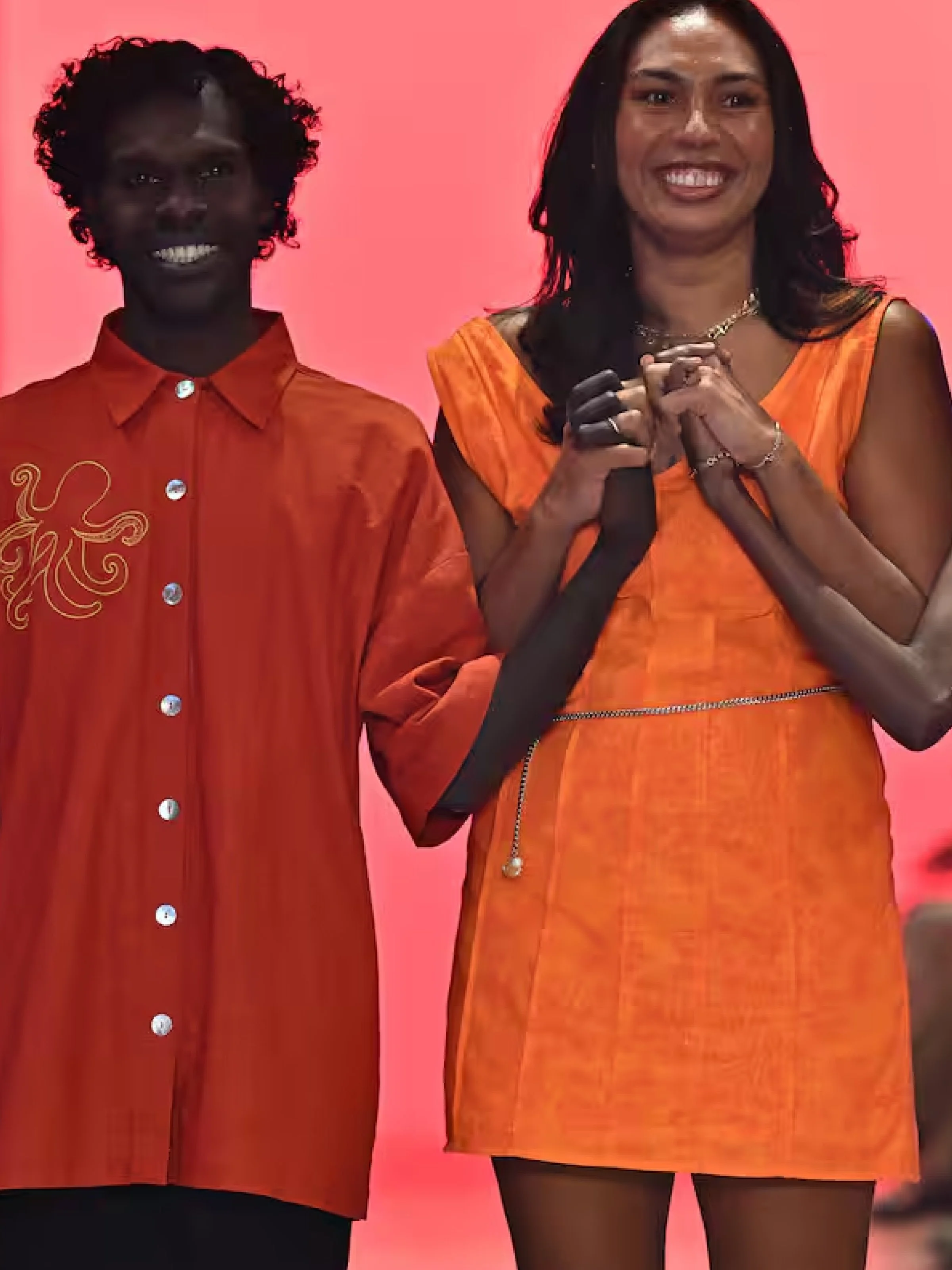 Two women dressed in orange, one with curly hair wearing a buttoned dress, and the other with straight hair in a sleeveless dress, standing against a pink background.
