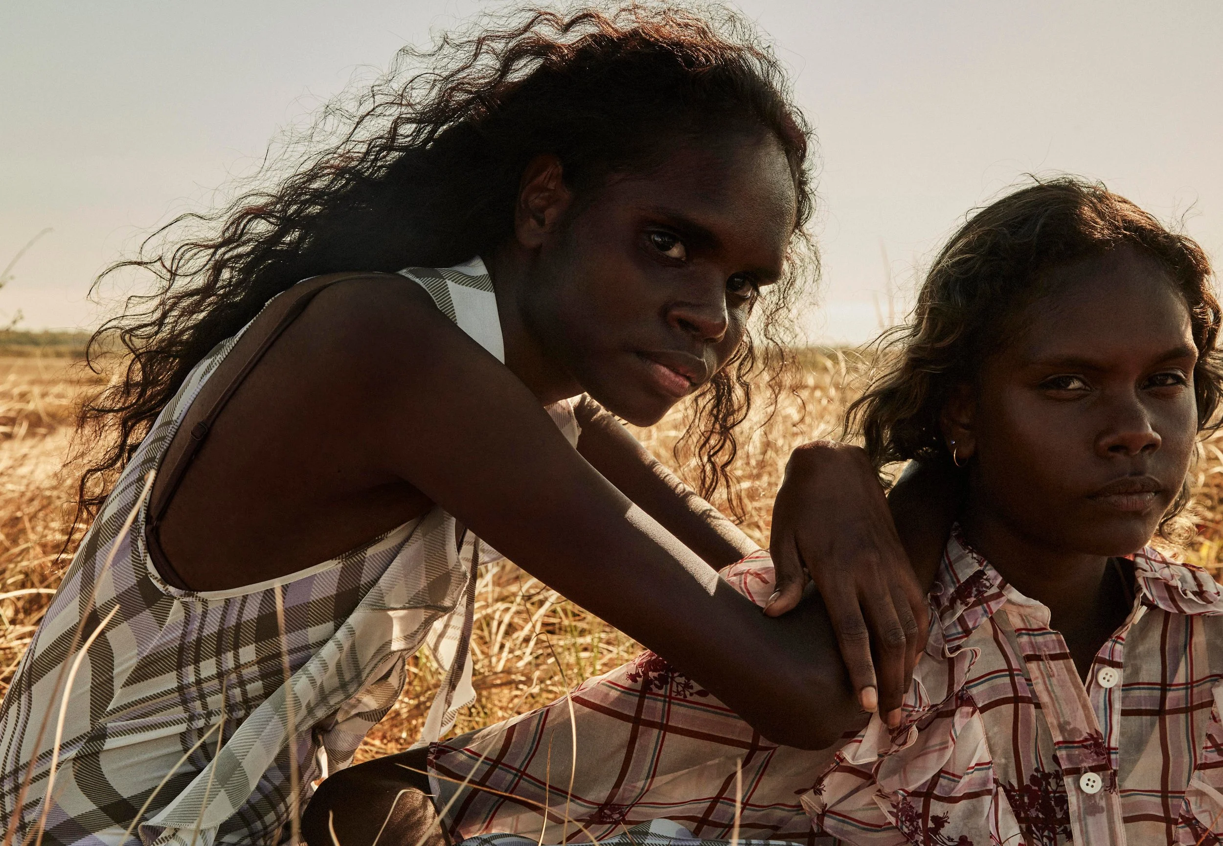 Two women with curly hair sitting in a field of dry grass during sunset or sunrise, looking at the camera, with serious expressions.