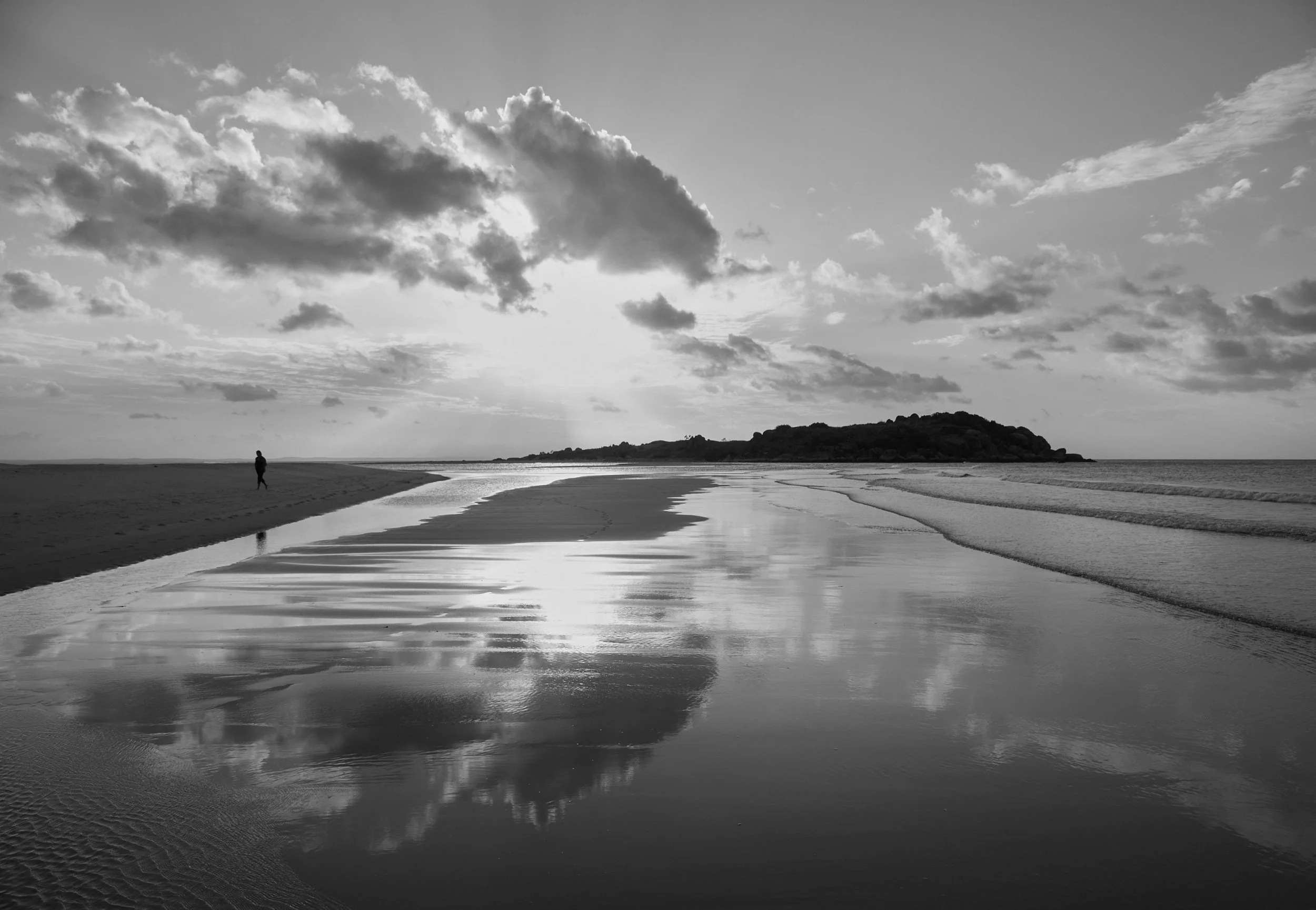 Black and white photograph of a person walking along a beach with water reflecting the cloudy sky, with an island or hill in the background.