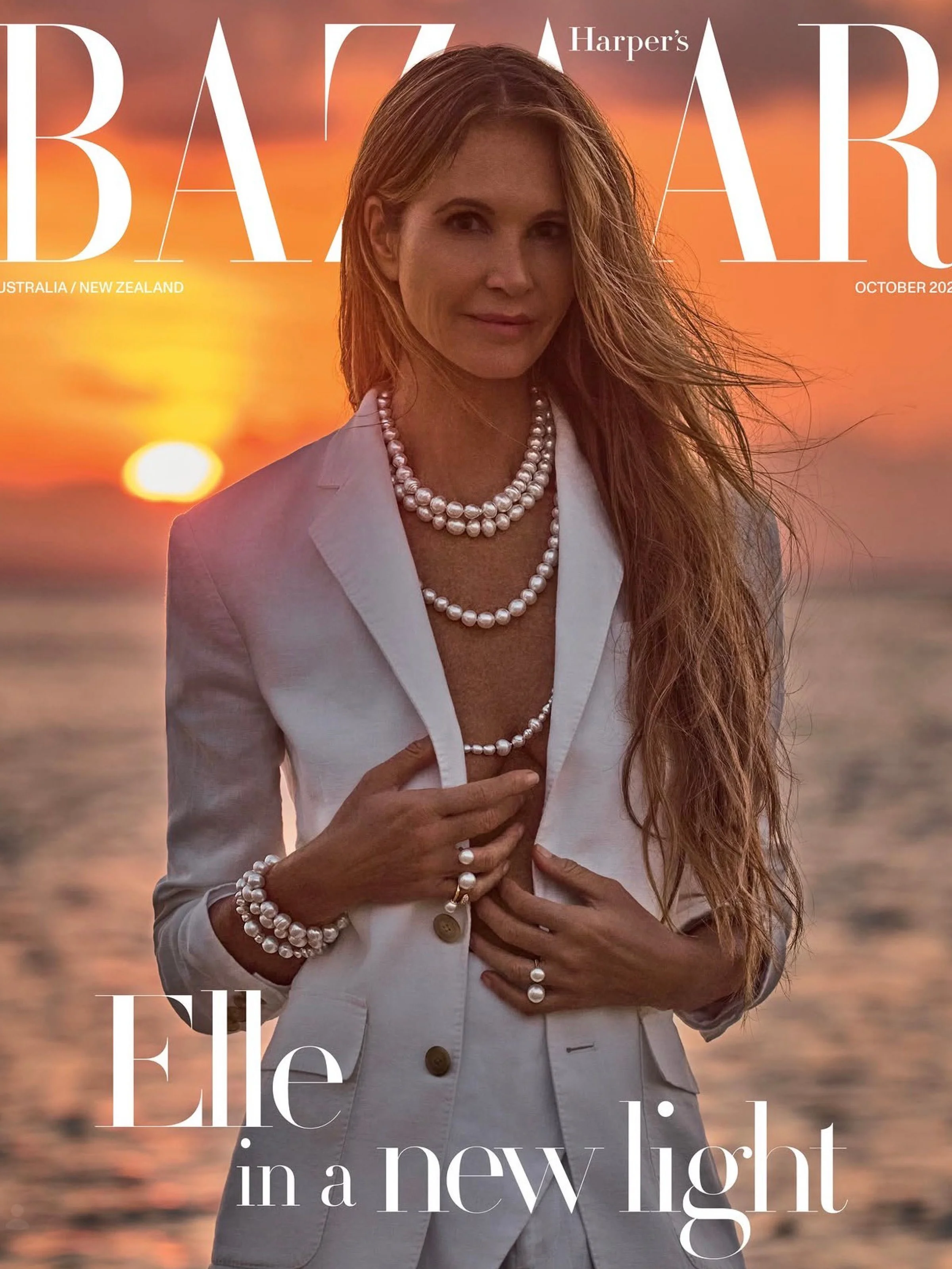 Woman with long wavy hair wearing white blazer and pearl jewelry standing on a beach at sunset.