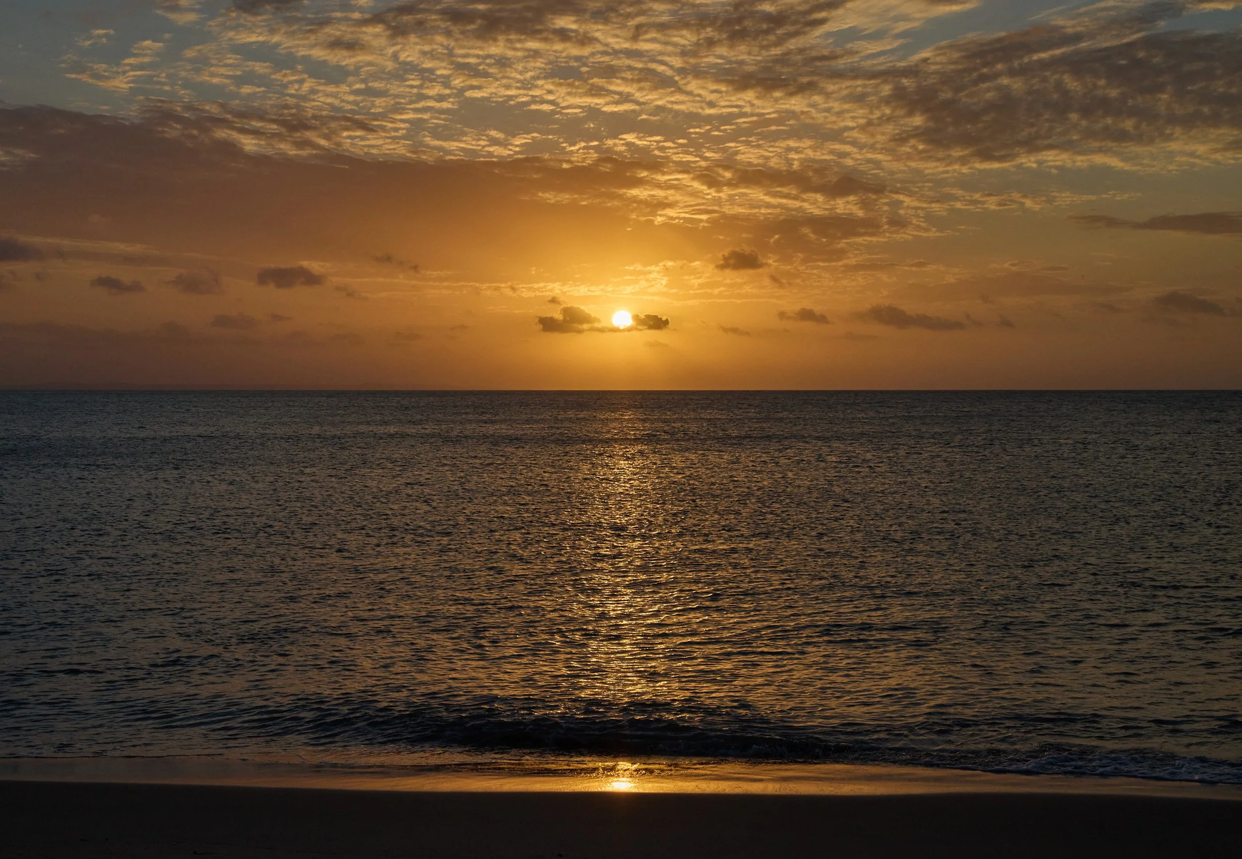 Sunset over the ocean with golden light, scattered clouds, and reflection on the water.