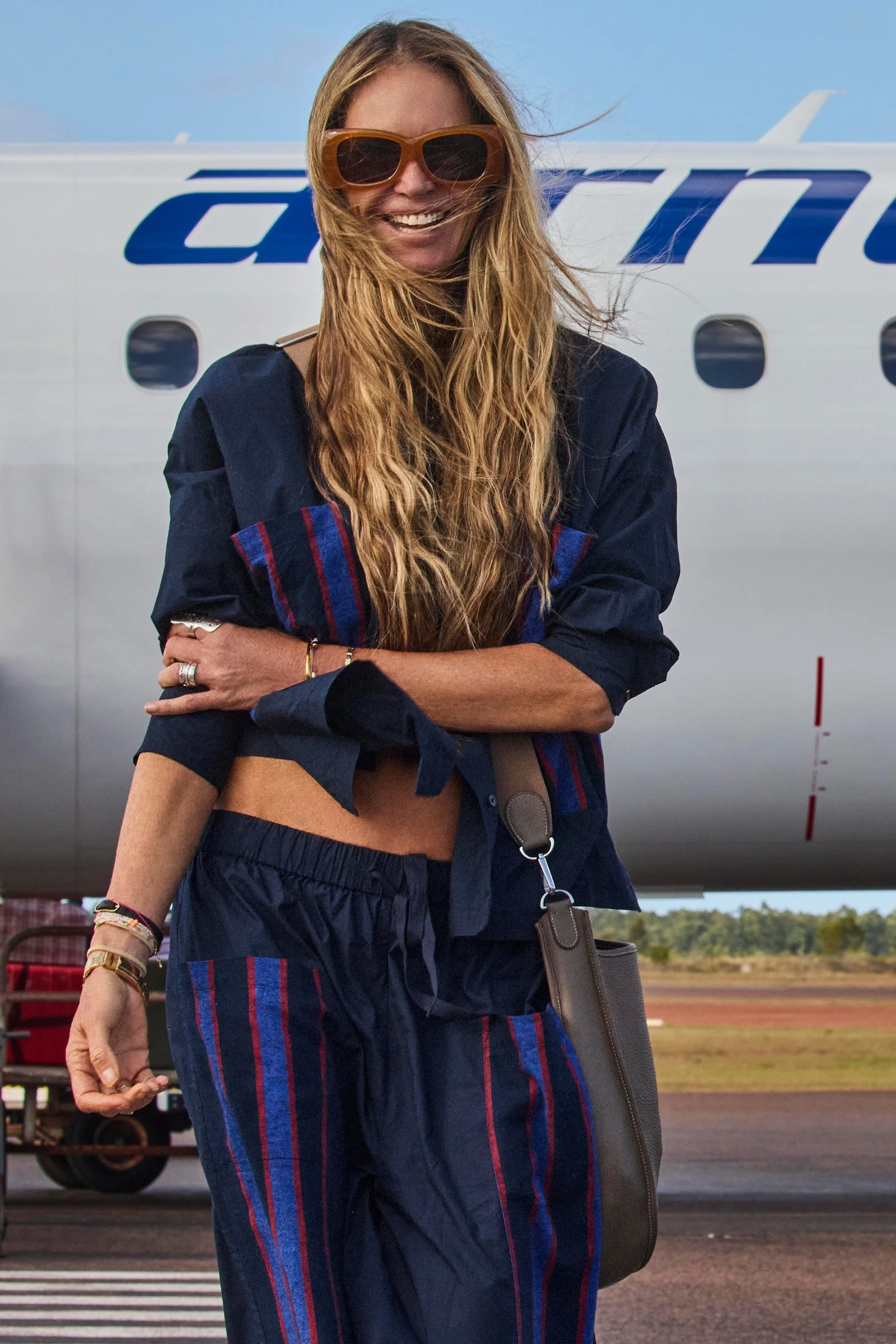 A woman smiling at an airport or airfield in front of an airplane, wearing sunglasses and casual clothing, with her long hair blowing in the wind.