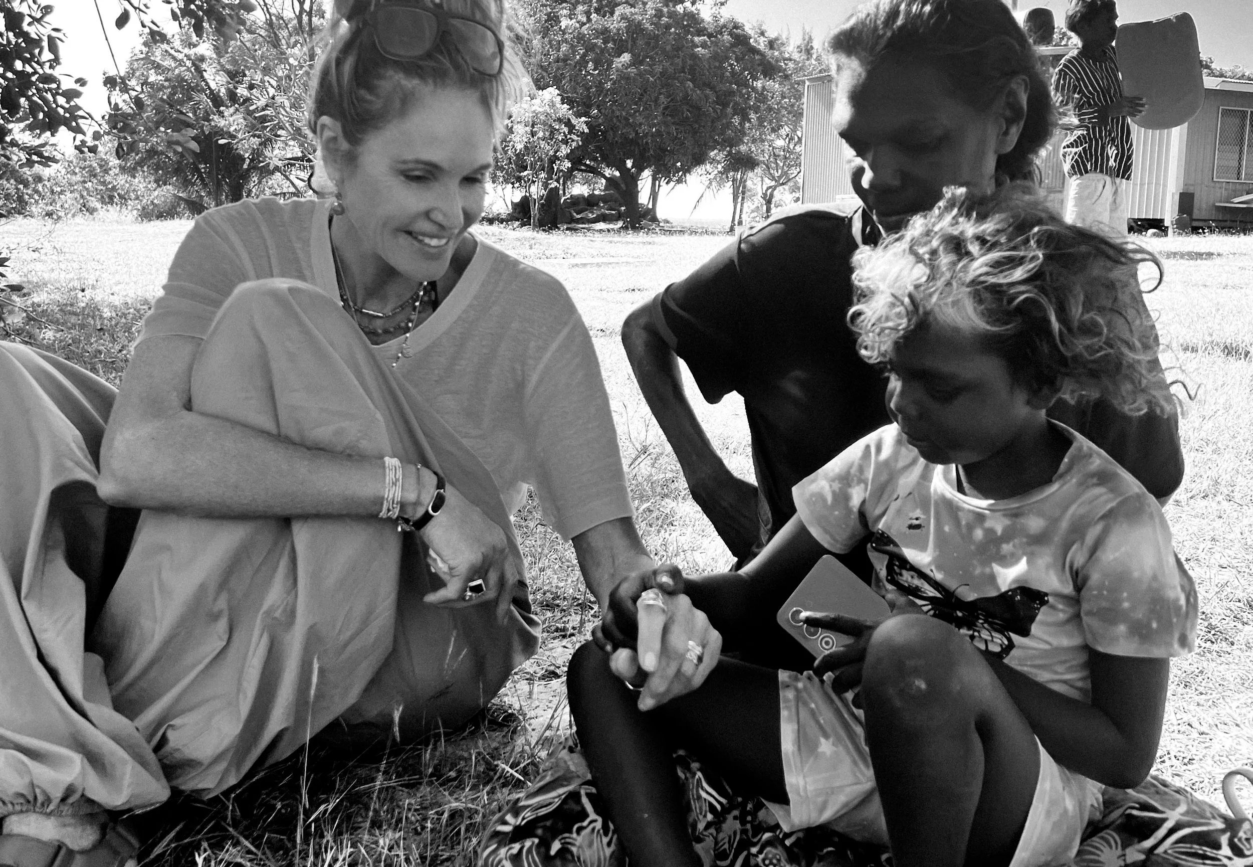 A group of people gathered outdoors in a grassy area with trees, some sitting and others standing, engaging in a social activity. The focus is on a woman with sunglasses on her head, smiling and interacting with two children, one of whom is holding a