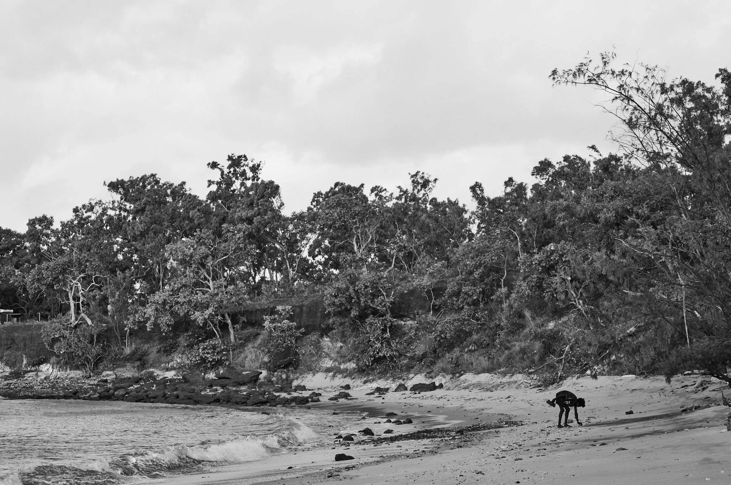A black and white photo of a beach shoreline with a person bending down near rocks and water. There are trees and hills in the background under an overcast sky.
