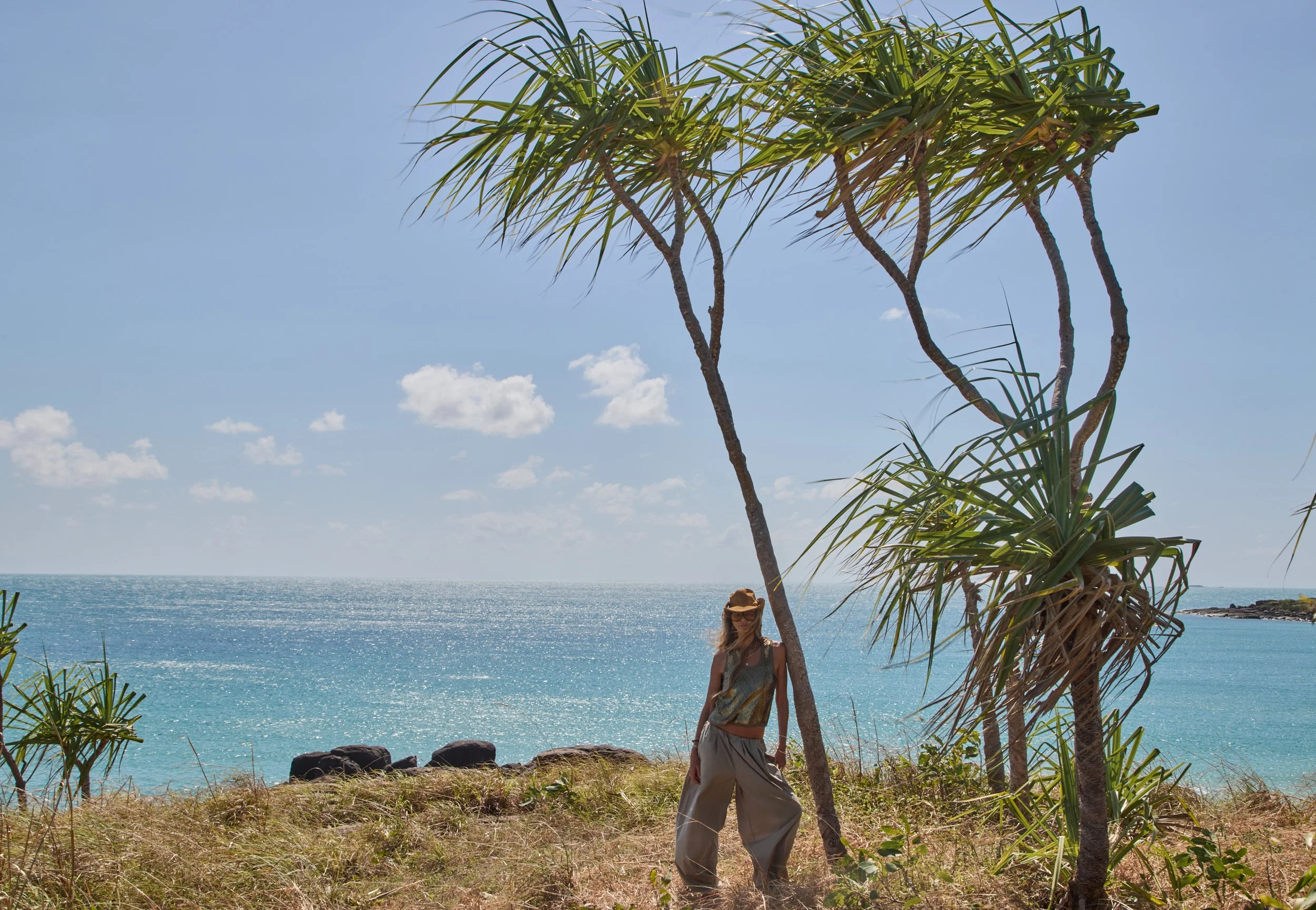 Woman standing under a small palm tree at a beach with turquoise water, rocky shoreline, and a distant island visible in the background. Clear blue sky with a few clouds.
