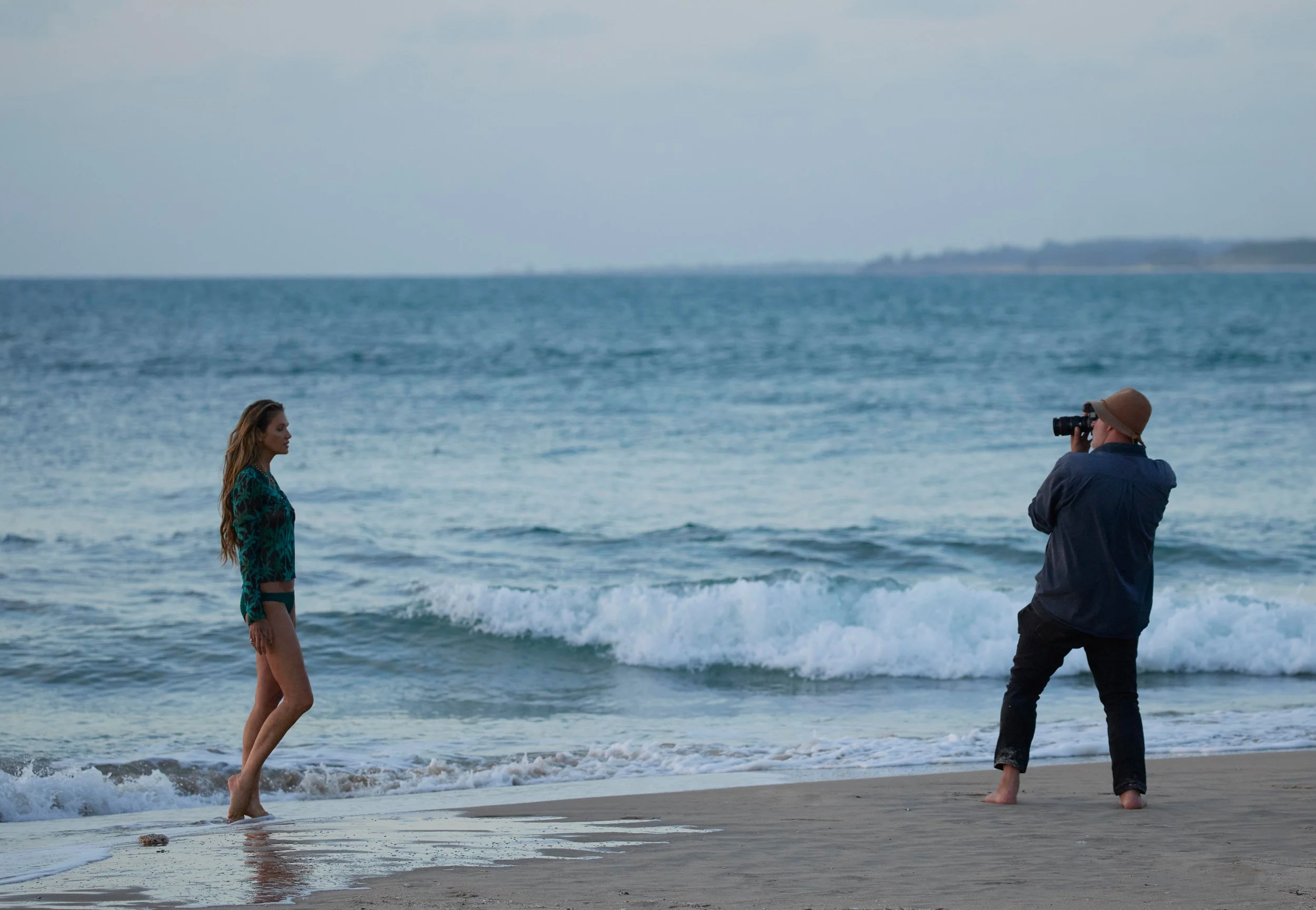 A woman with long wavy hair standing at the shoreline facing the ocean, wearing a patterned dress, while a man with a hat takes a photo of her with a camera by the water.