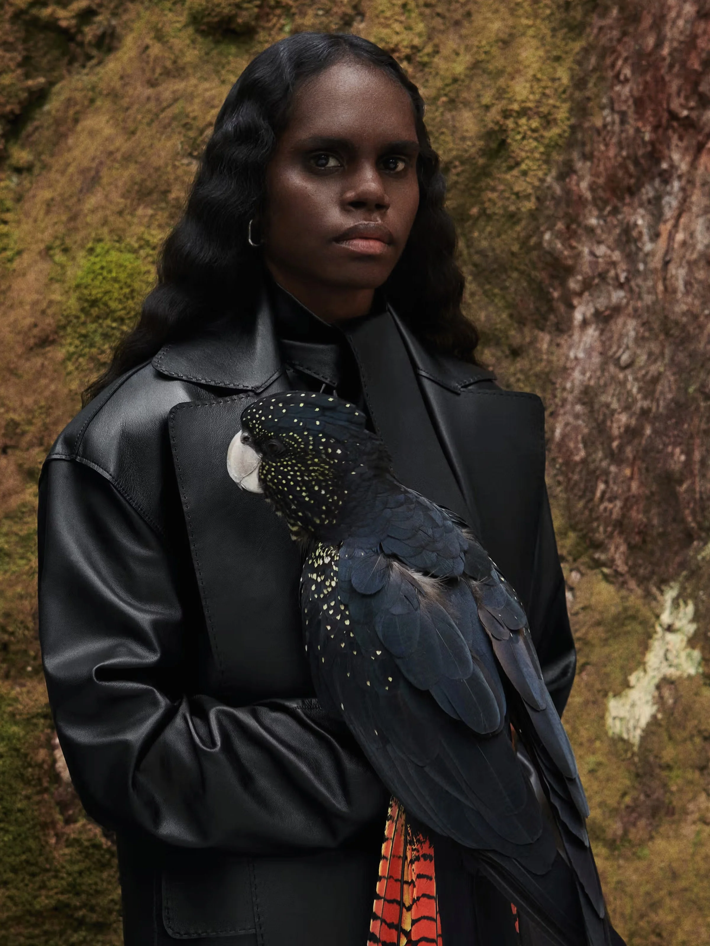 A woman with dark skin and long wavy black hair standing outdoors against a textured rocky background. She is wearing a black leather jacket and holding a large black bird with spotted yellow and green feathers, likely a bird of prey.