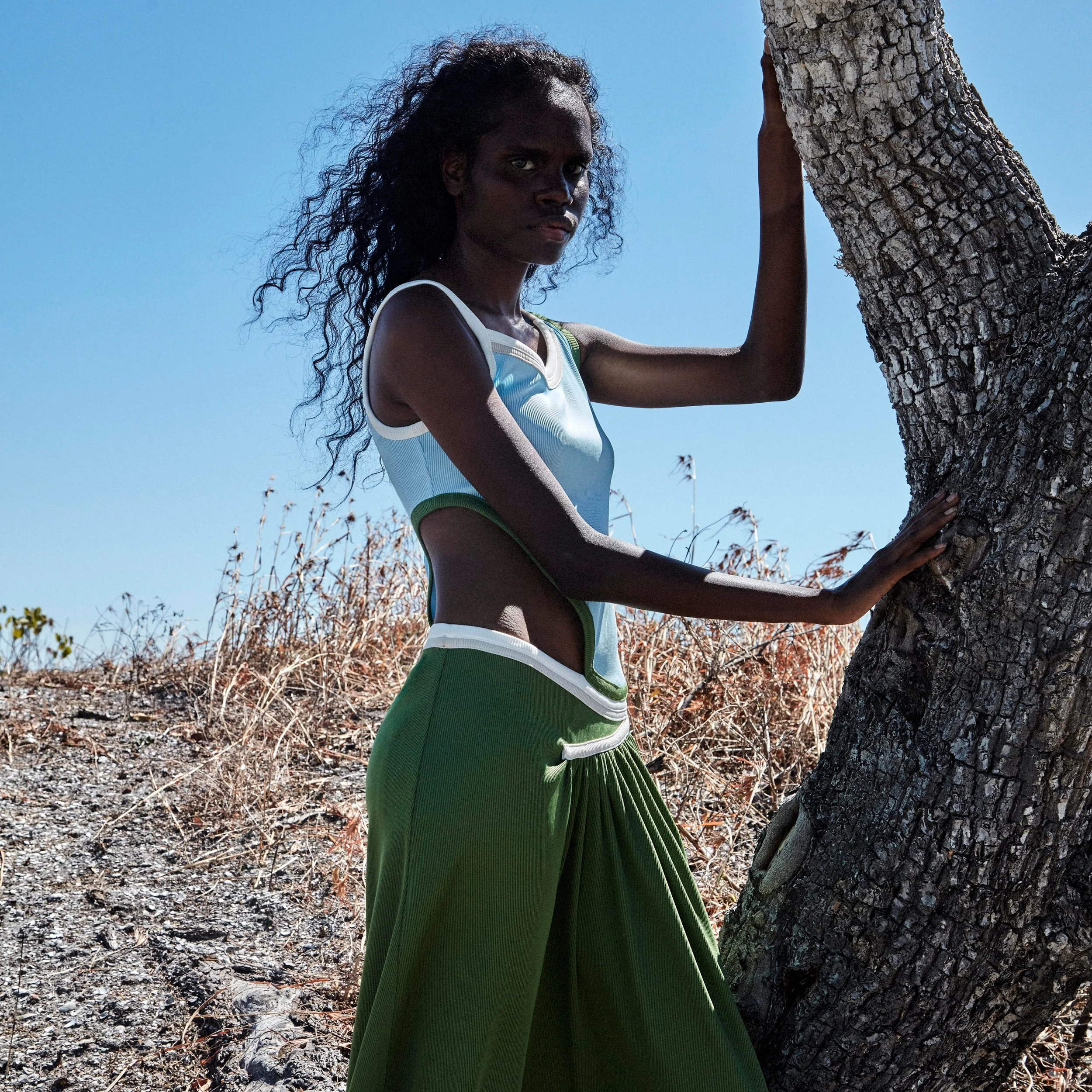 Tarlisa Gaykamangu standing beside a tree in East Arnhem Land, wearing a green skirt and looking toward the camera.