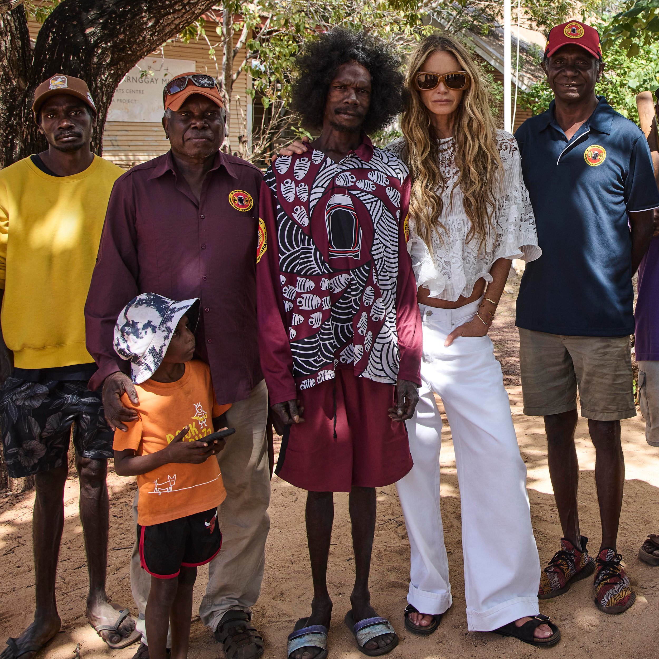 Group of five people standing outdoors under a tree, with two women and three men, some wearing sunglasses and casual attire, a mix of Indigenous and non-Indigenous individuals, with shoes placed on the ground in front of them.